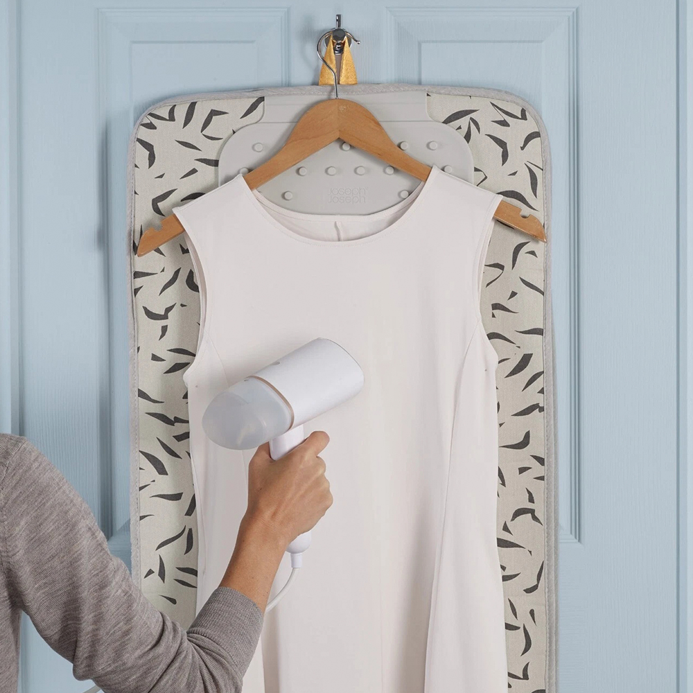Una persona plancha un vestido blanco en una percha, colgado en una puerta con una tabla de planchar estampada detrás.