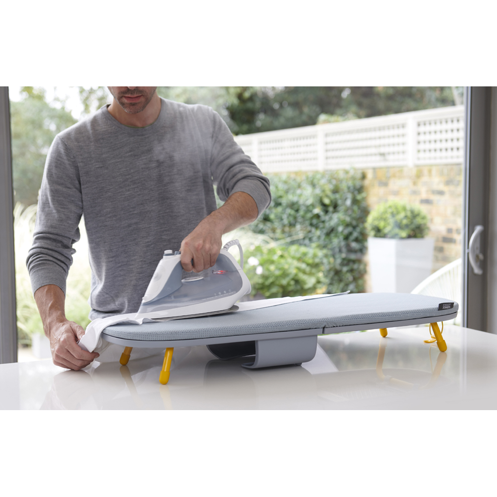 Un hombre plancha ropa en una tabla de planchar pequeña. La tabla está sobre una mesa blanca frente a una ventana. La plancha es blanca y azul.