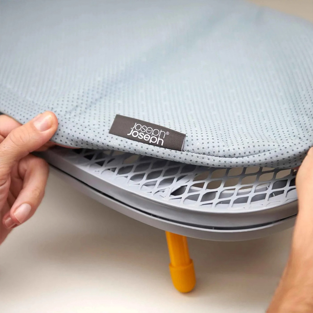 Manos sosteniendo una funda gris de tabla de planchar con perforaciones. Se ven un marco gris, una pata amarilla y el logo 'Joseph Joseph'.