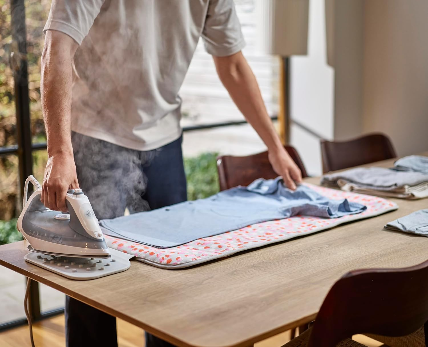 Una persona plancha una camisa azul sobre una mesa. Una plancha emite vapor. Hay otras camisas dobladas sobre la mesa.