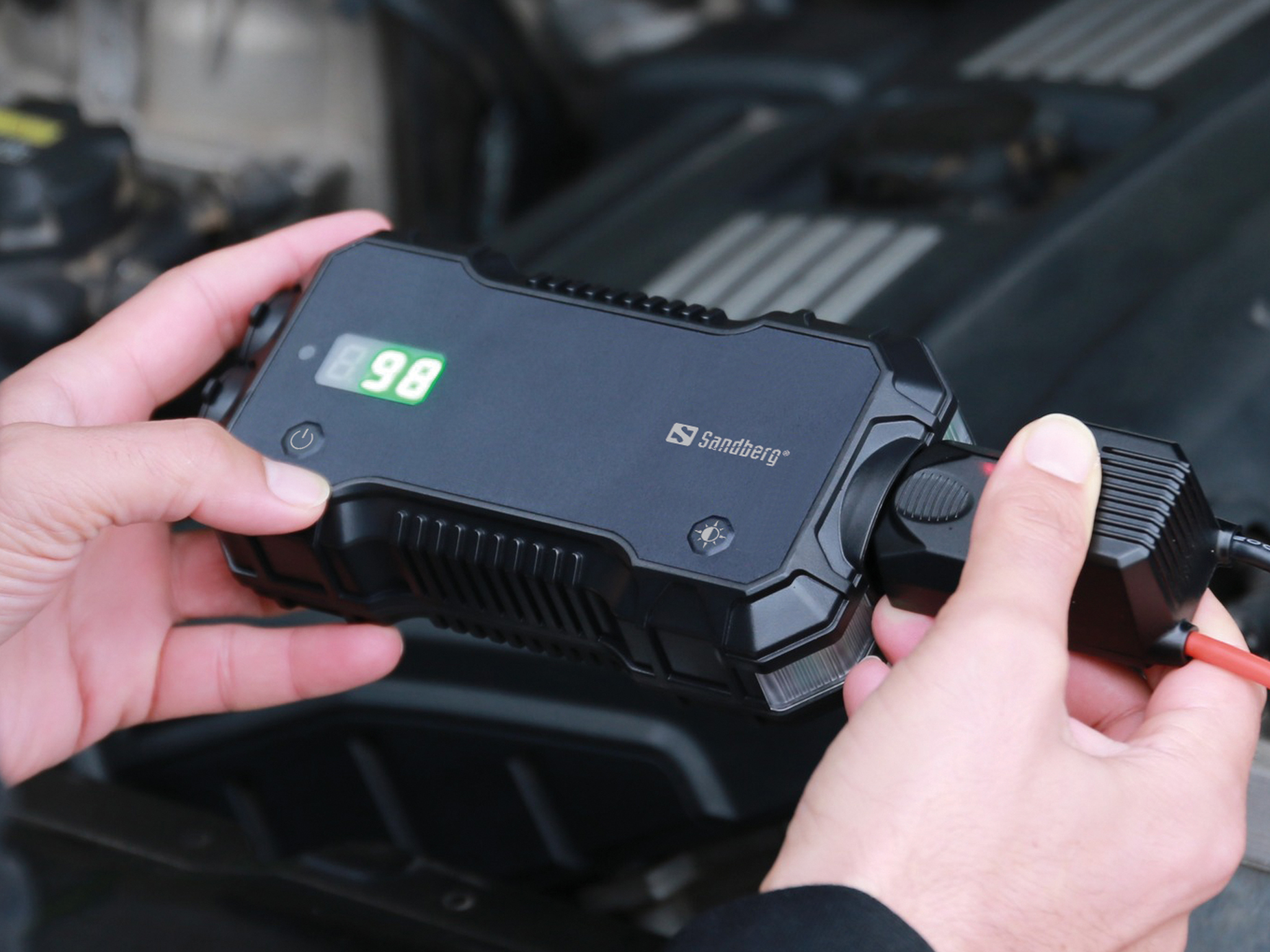 Le mani tengono un avviatore Sandberg nero, che mostra la carica al 98%. Il dispositivo è collegato a un motore di un'auto.