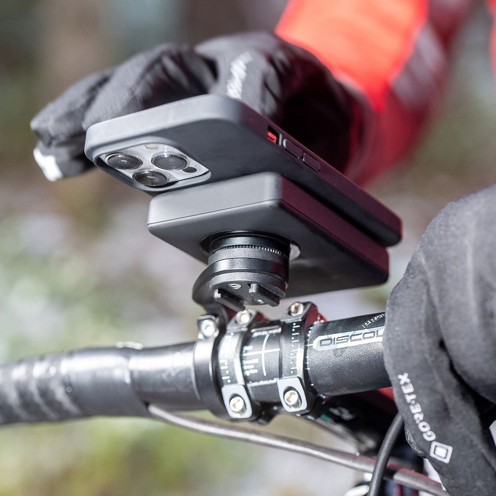 Un telefono montato sul manubrio di una bicicletta. Mani con guanti tengono il telefono. Lo sfondo è sfocato.