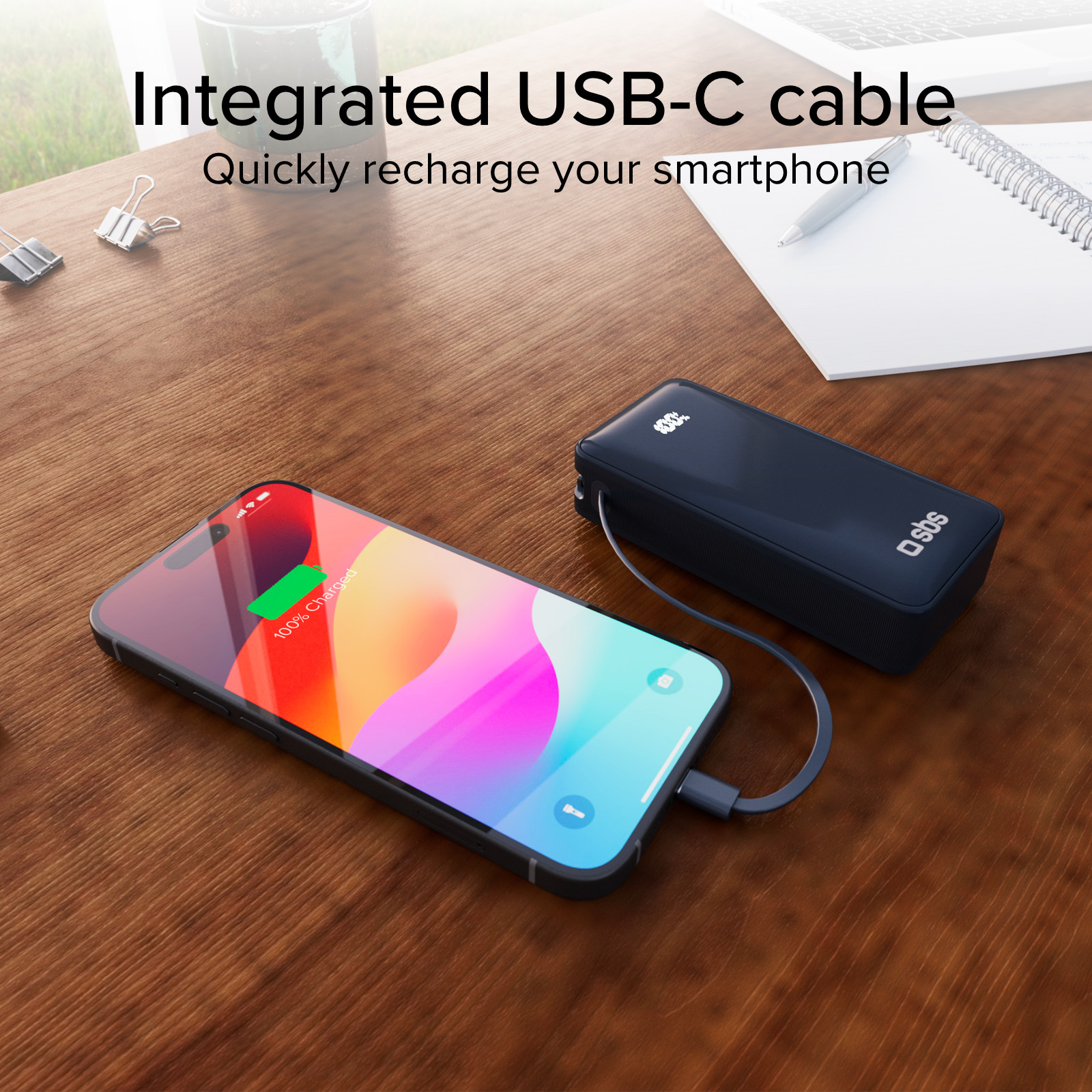 Smartphone in carica con cavo USB-C collegato a un power bank, entrambi su un tavolo di legno. Il telefono mostra 100% carica.