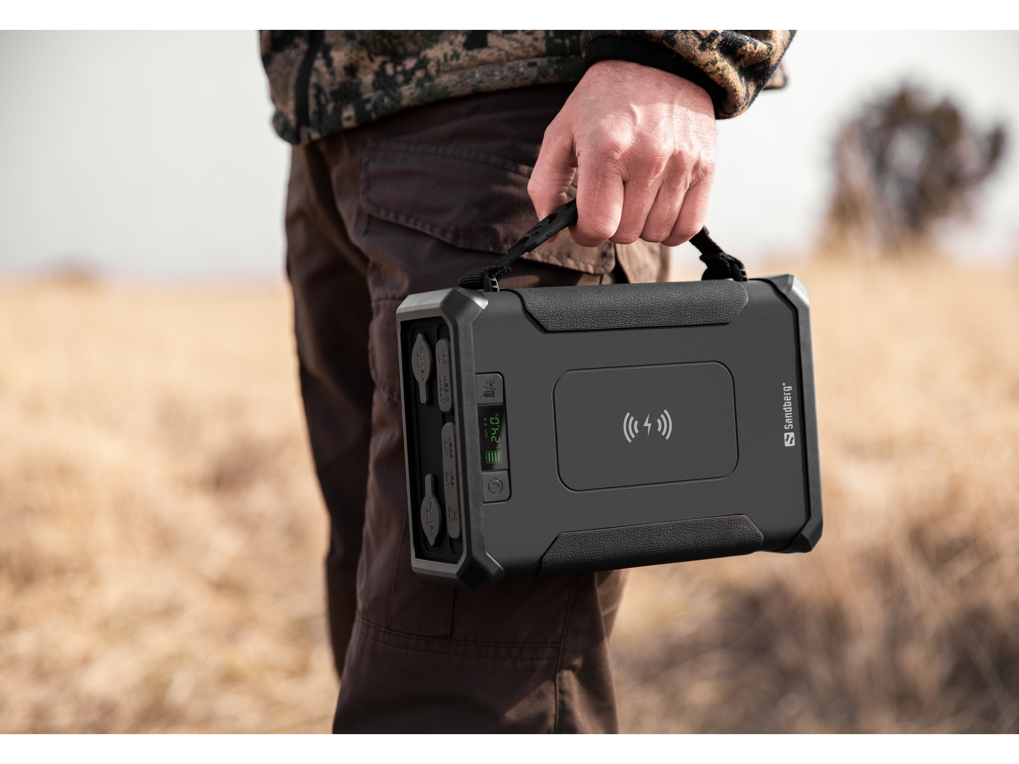Una persona tiene in mano un power bank nero con manico. Lo sfondo è un campo con erba secca.