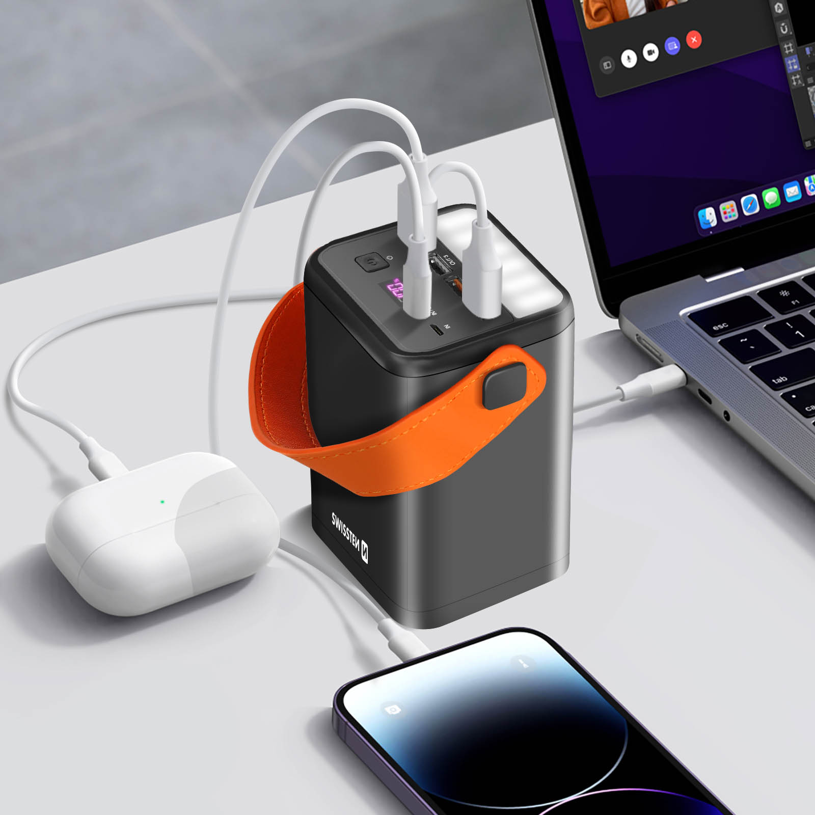 Un power bank nero con manico arancione. I cavi si collegano a telefono, laptop e auricolari, tutto su una superficie bianca.