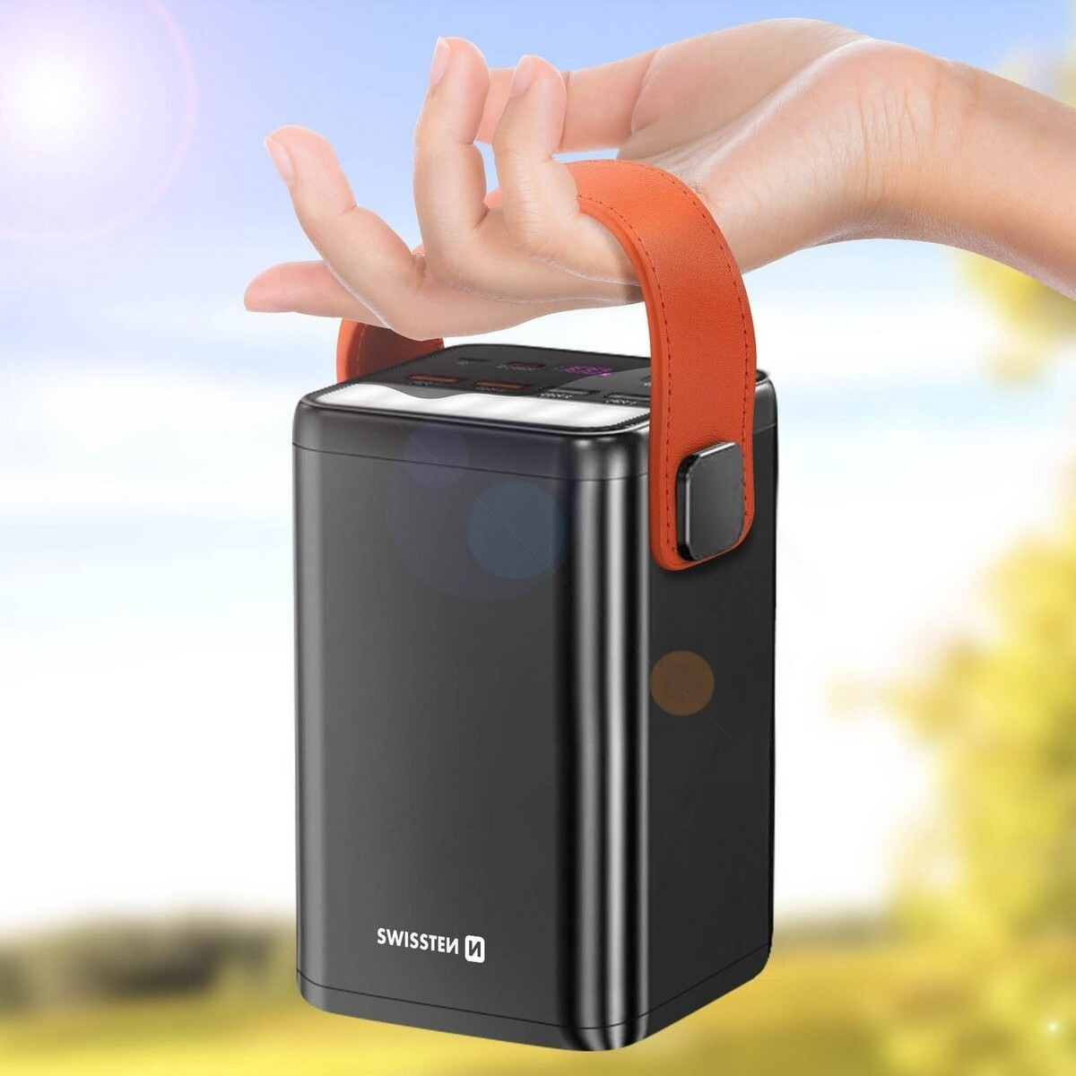 Una mano tiene in mano un power bank nero con un manico arancione. Lo sfondo è sfocato, suggerendo un ambiente esterno.