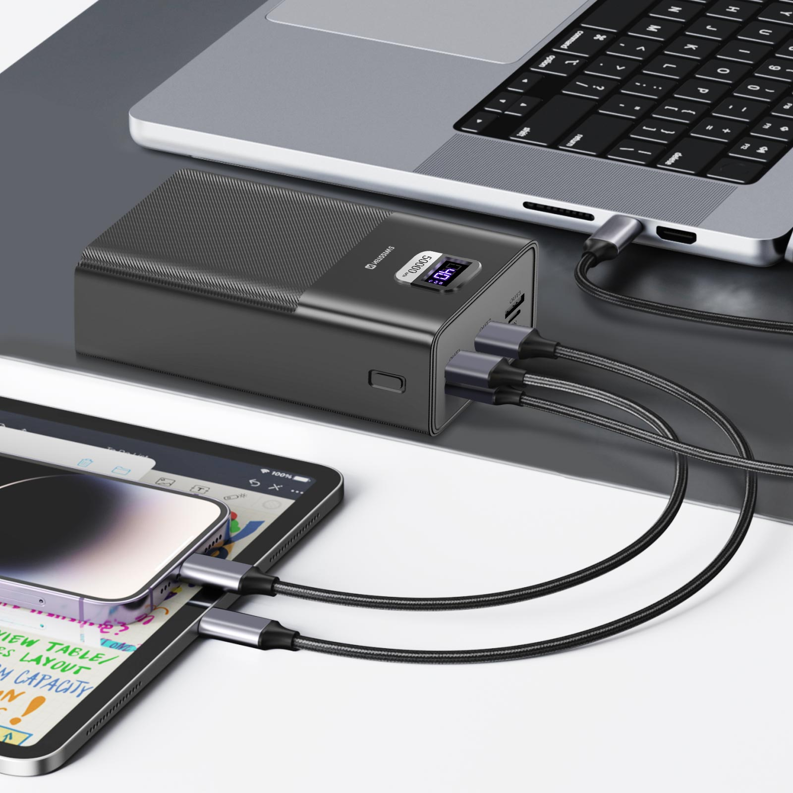 Un power bank nero con cavi collegati a un laptop, un tablet e un telefono, il tutto su una superficie bianca.
