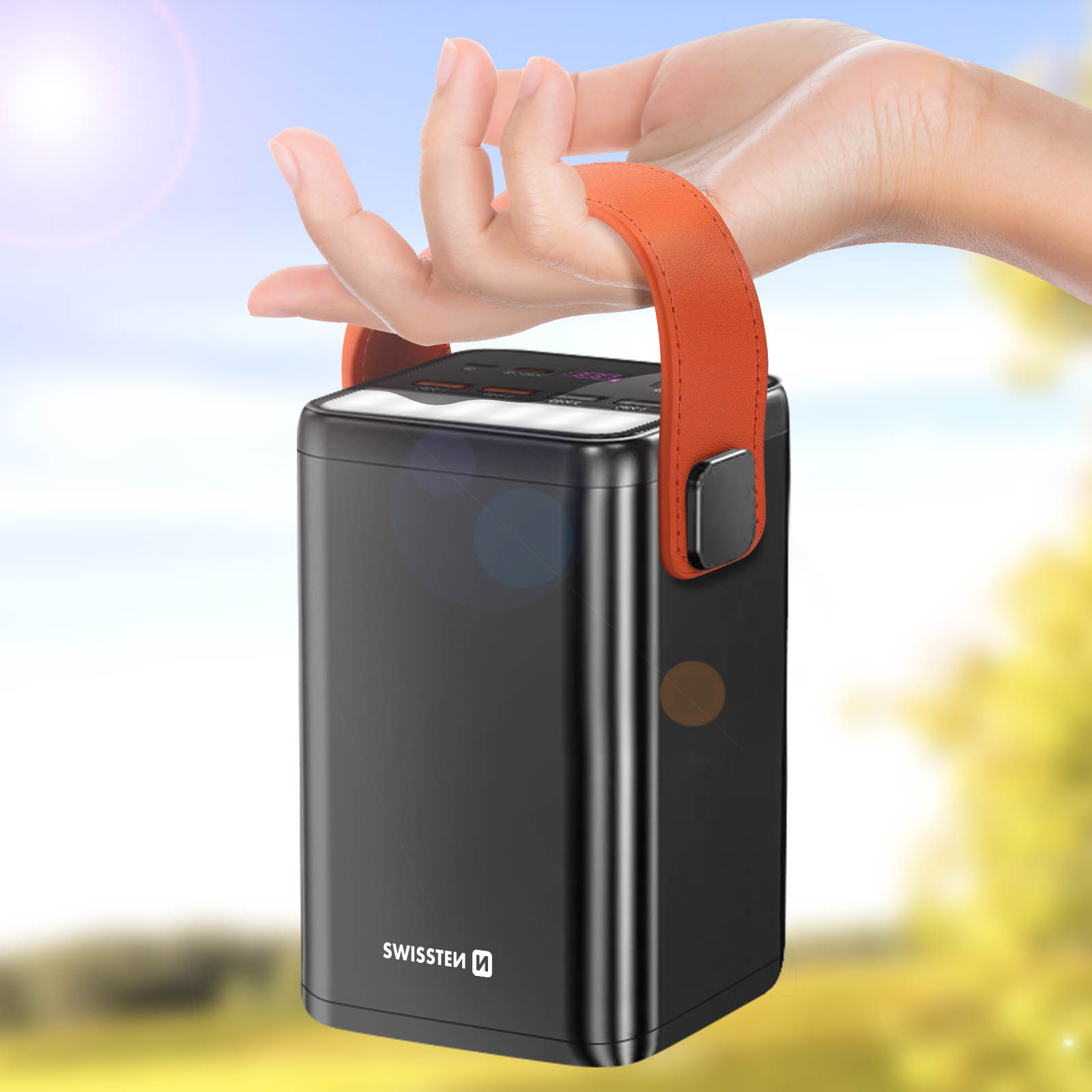 Una mano tiene un power bank nero con un cinturino arancione. Lo sfondo è una scena sfocata all'aperto con la luce solare.