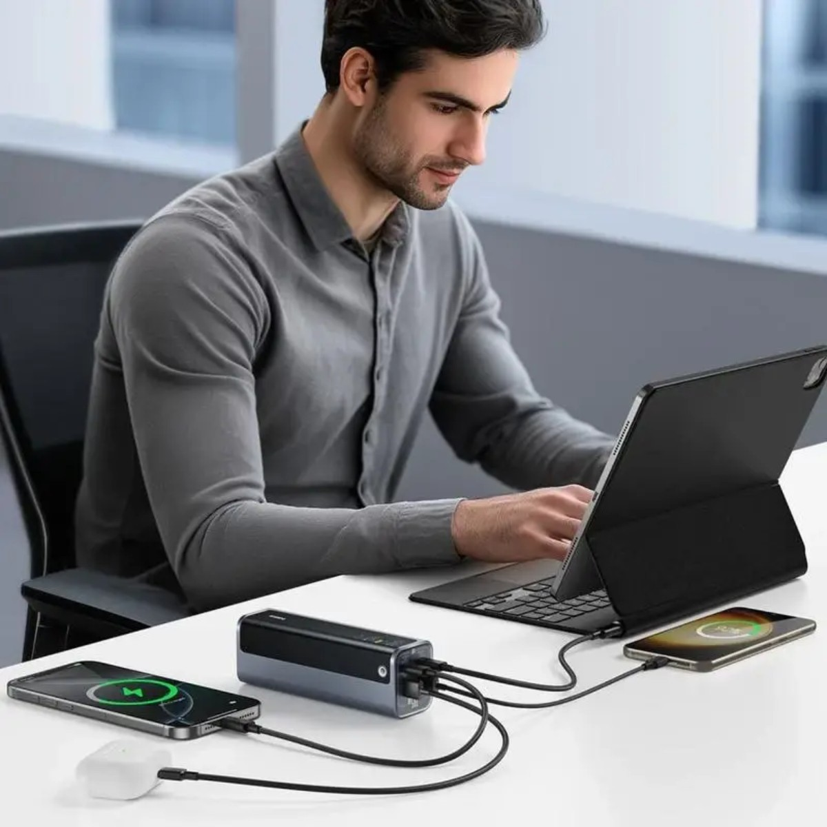 Un uomo usa un laptop, caricando più dispositivi con un power bank. Telefoni, tablet e auricolari sono collegati. La scena è al chiuso.