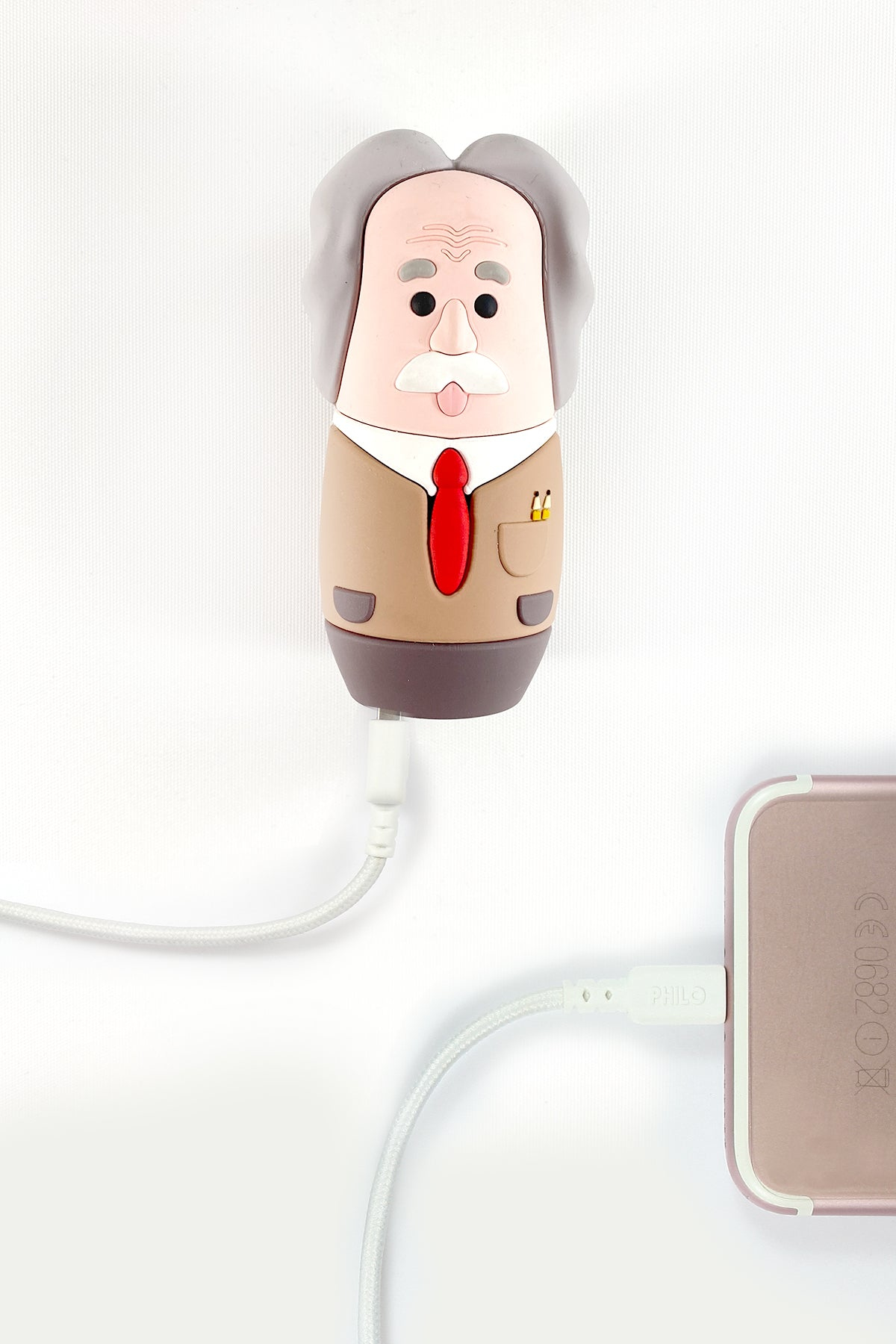 Un caricabatterie a forma di Einstein è collegato a un telefono. Il caricabatterie è bianco e il telefono è oro rosa. Sfondo bianco.