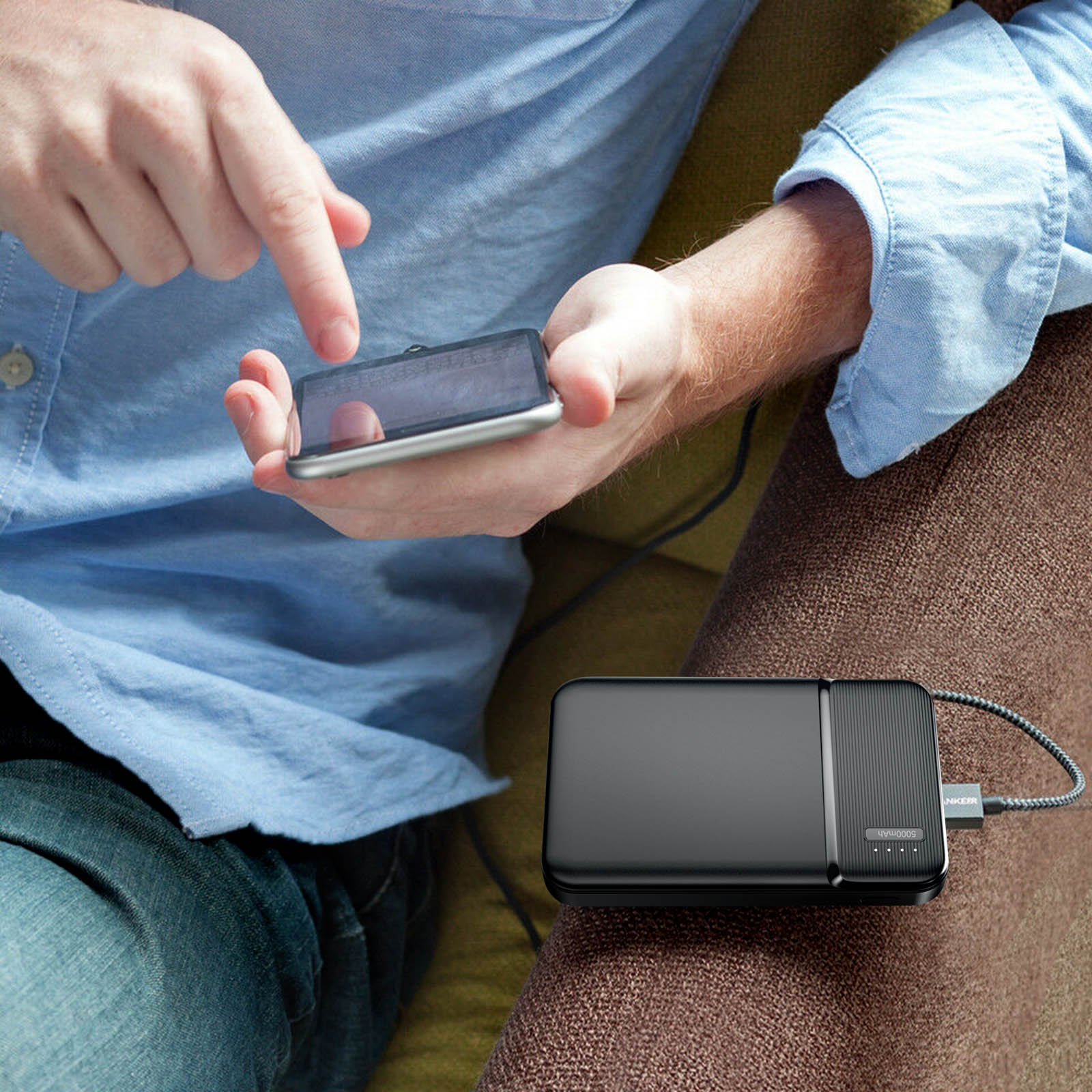 Una persona usa un telefono con un power bank collegato. Indossa una camicia blu. La scena è al chiuso, su un divano.