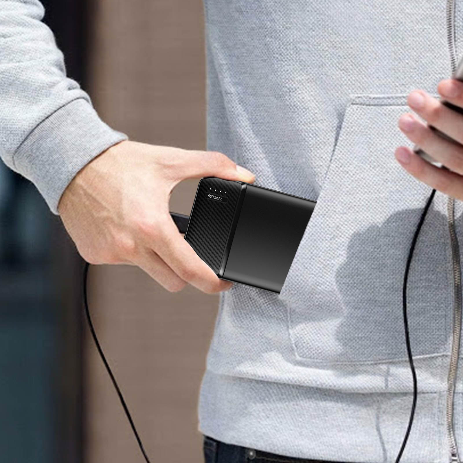 Una persona tiene in mano un power bank nero, parzialmente in tasca. La persona indossa una felpa con cappuccio grigia. Un telefono è tenuto.
