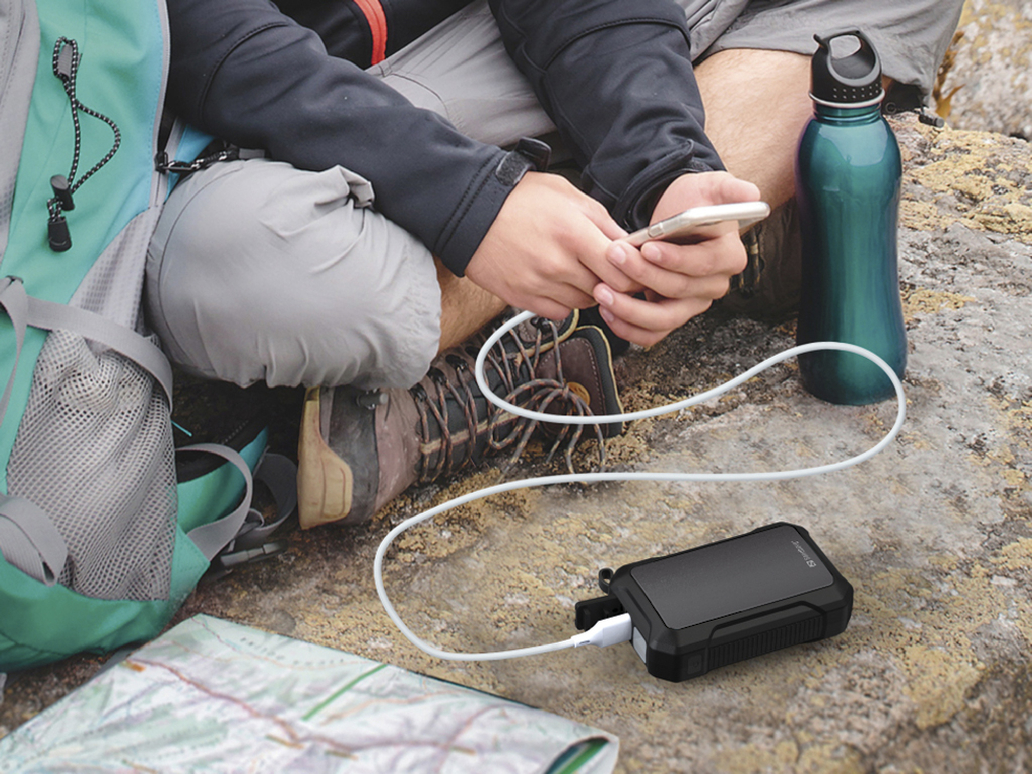 Een persoon gebruikt een telefoon, verbonden met een powerbank via een witte kabel. Een rugzak, kaart en waterfles zijn ook aanwezig.