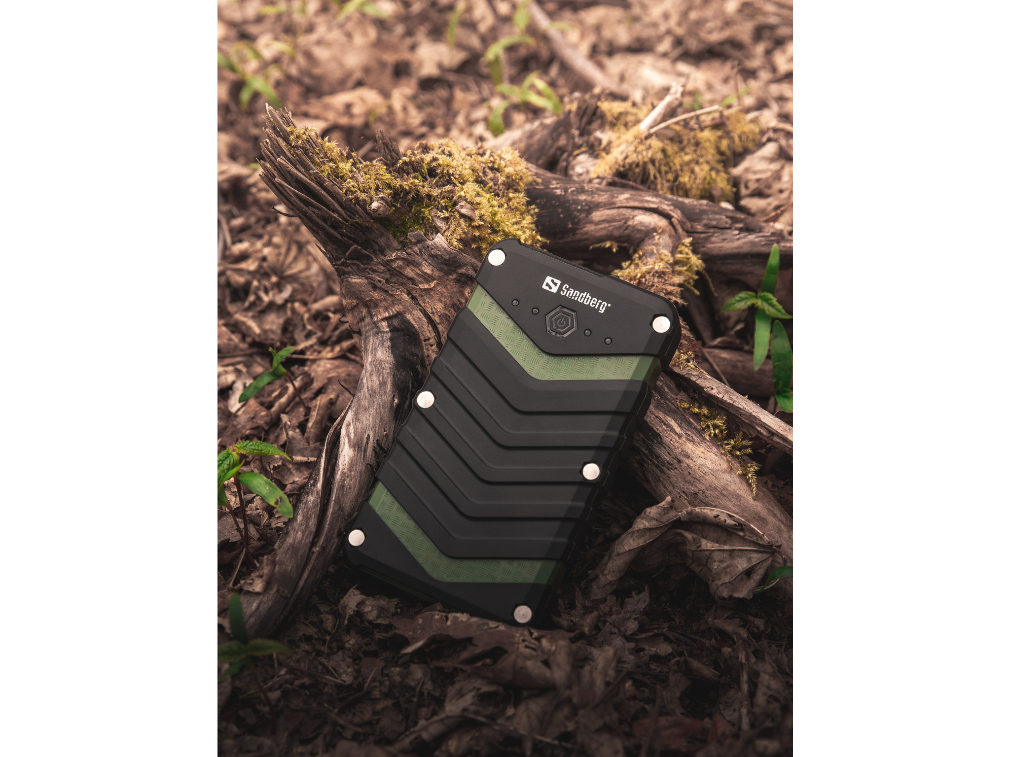 Power bank Sandberg nero e verde su un tronco di legno con muschio, circondato da foglie e piante.