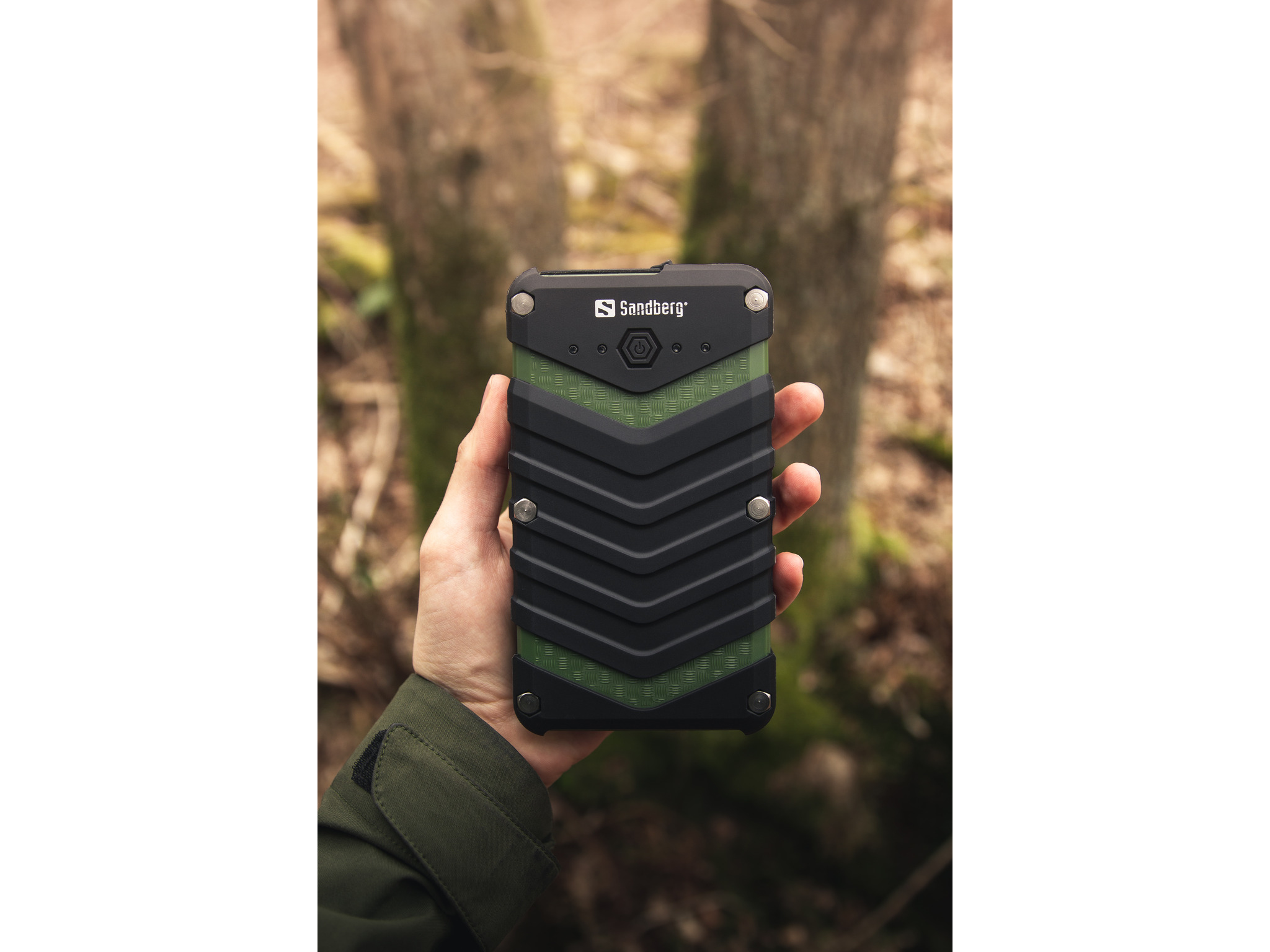 Una mano tiene un power bank Sandberg. È nero e verde, con un design a zig-zag. Gli alberi sono sfocati sullo sfondo.