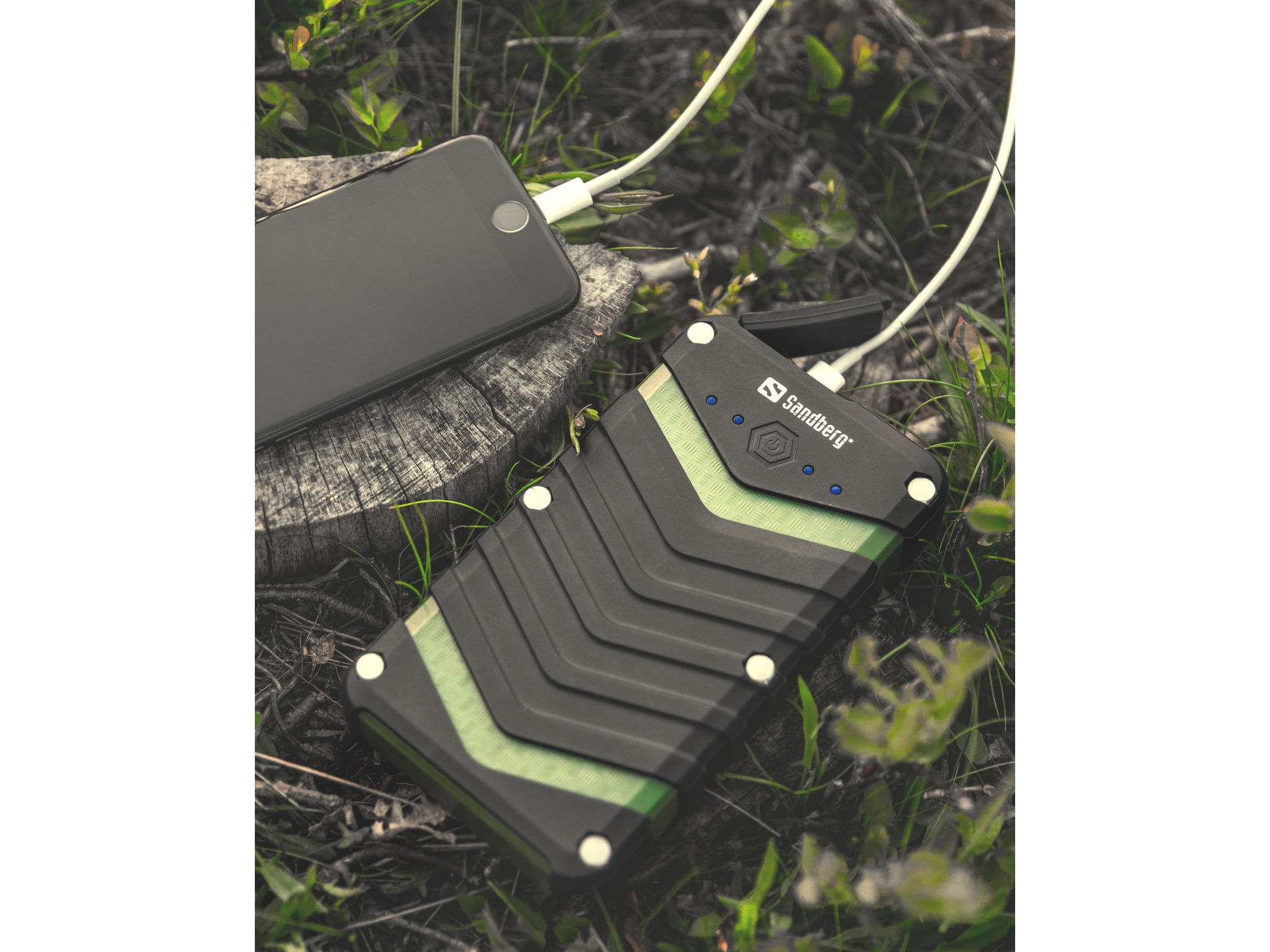 Un telefono e un power bank nero e verde sono collegati con un cavo bianco. Sono su una superficie di legno circondata dall'erba.