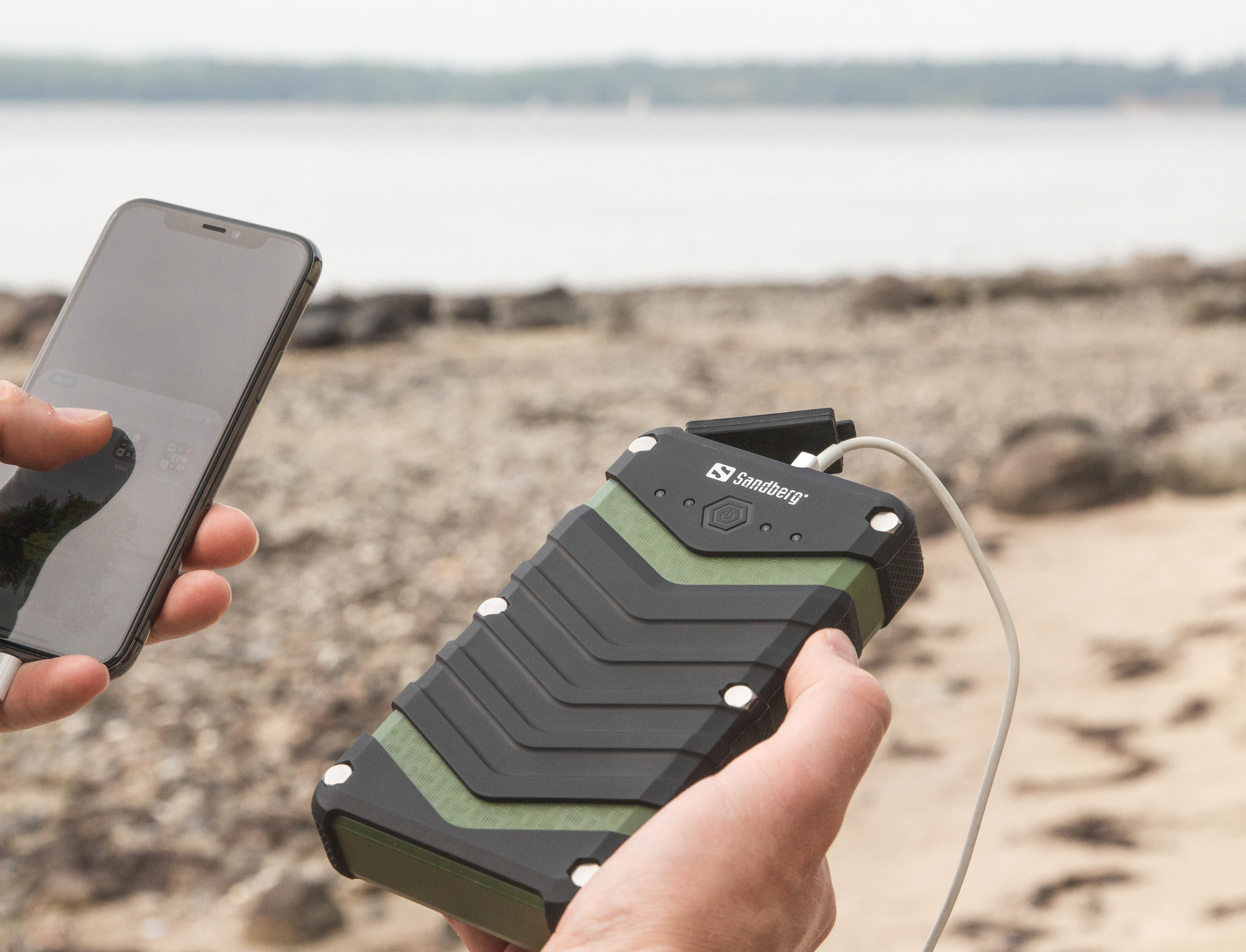 Una persona tiene in mano un telefono nero e un power bank nero e verde. Lo sfondo mostra una spiaggia e uno specchio d'acqua.