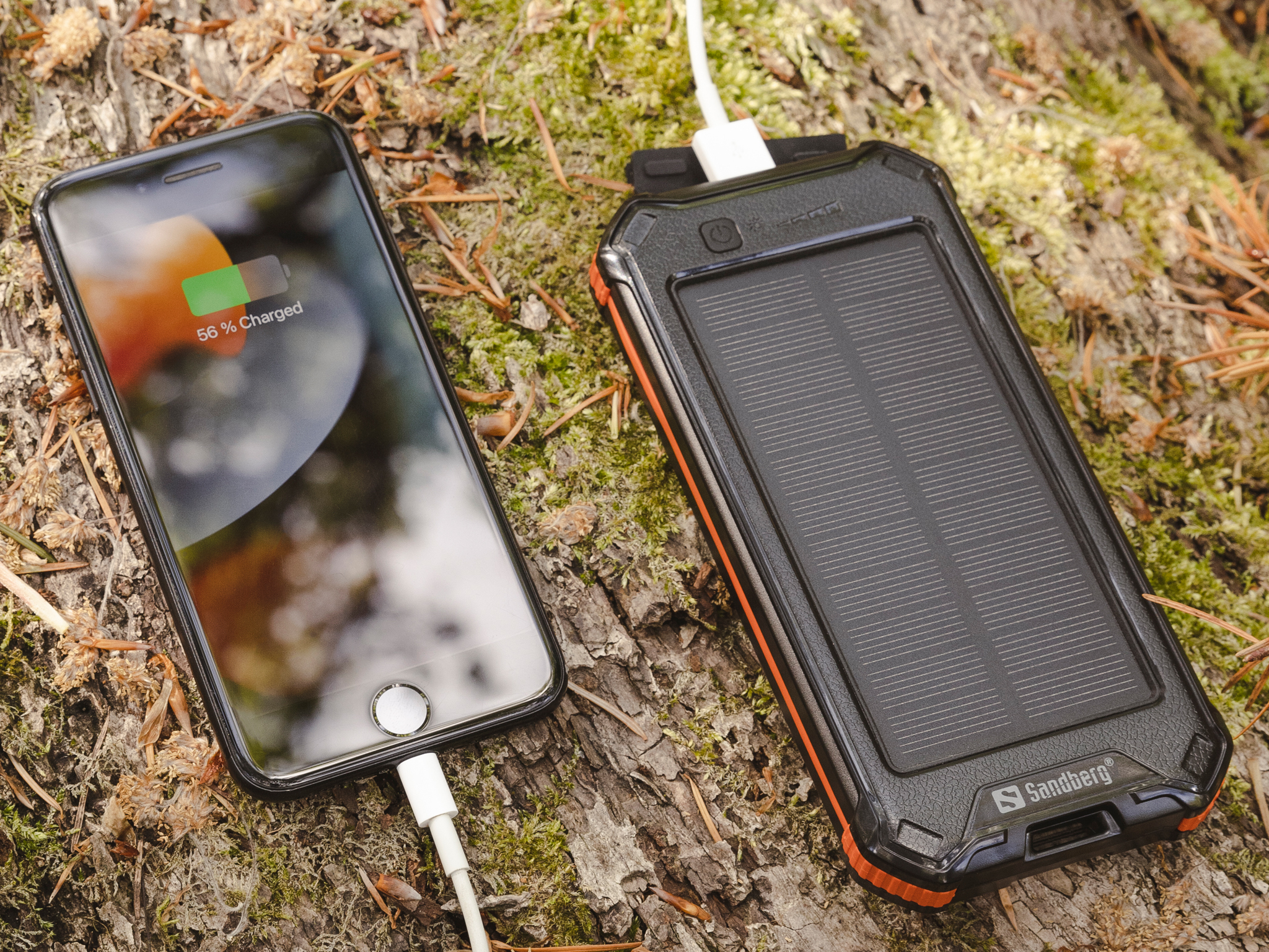 Un telefono in carica, mostra il 56% di batteria. Collegato a un caricabatterie solare portatile nero e arancione, posto su legno e muschio.