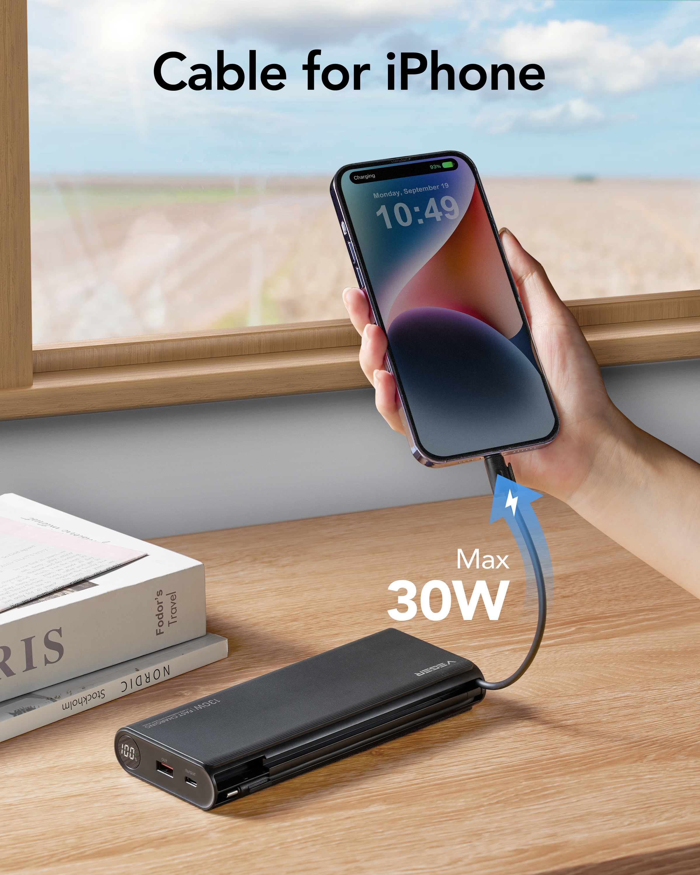 Un iPhone è in carica tramite un cavo collegato a un power bank, un libro e una finestra sono visibili sullo sfondo.