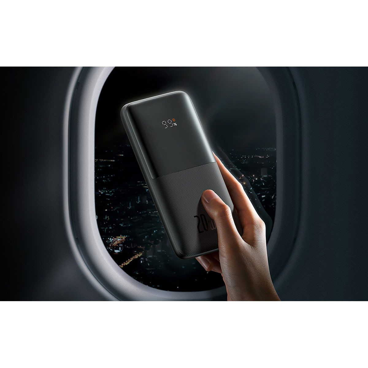 Una mano tiene un power bank nero vicino al finestrino di un aereo, mostrando il 99% di carica. Luci della città visibili sullo sfondo.