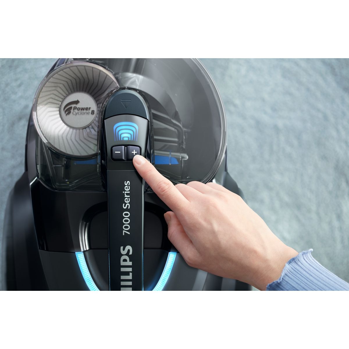 Una mano presiona el botón '+' en una aspiradora Philips. La aspiradora es negra, con luces azules. El fondo es una alfombra gris.