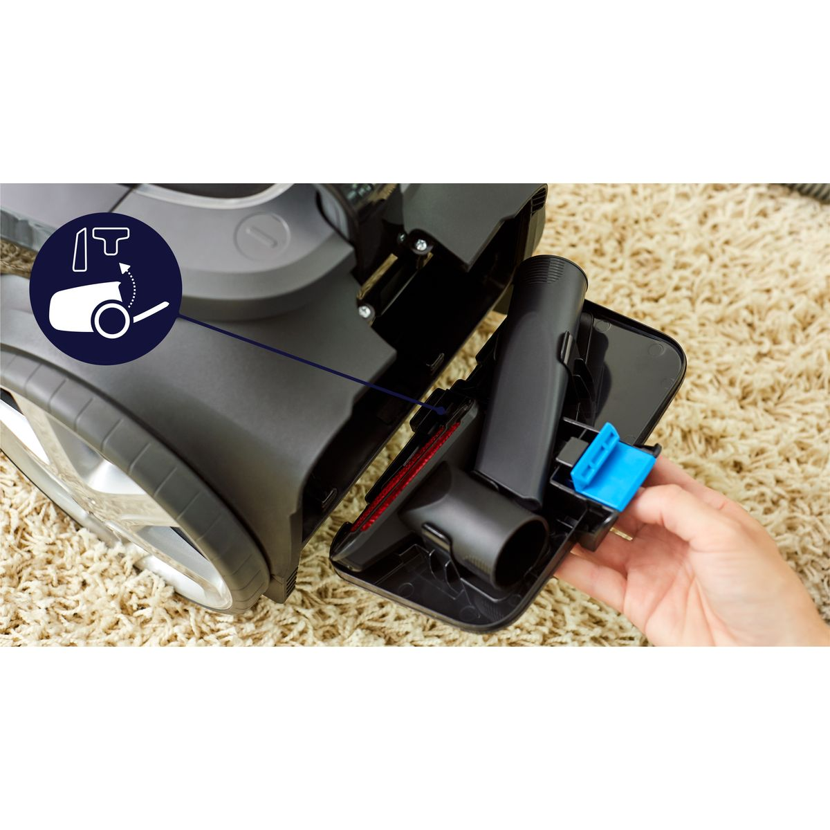 Odkurzacz z otwartą komorą, pokazujący akcesoria. Ręka trzyma niebieski element. Tło to beżowy dywan.