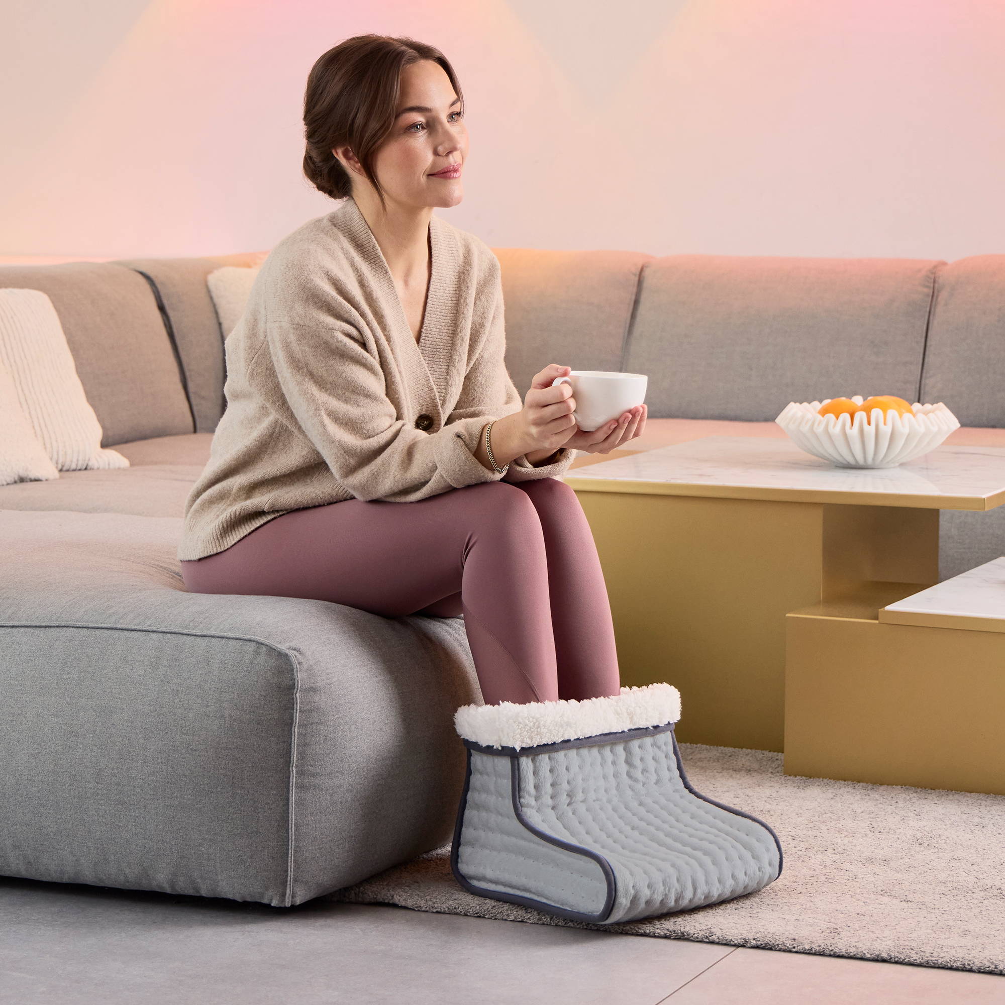 Mujer en leggings y suéter sentada con un calentador de pies, sosteniendo una taza. Un sofá y una mesa están al fondo.
