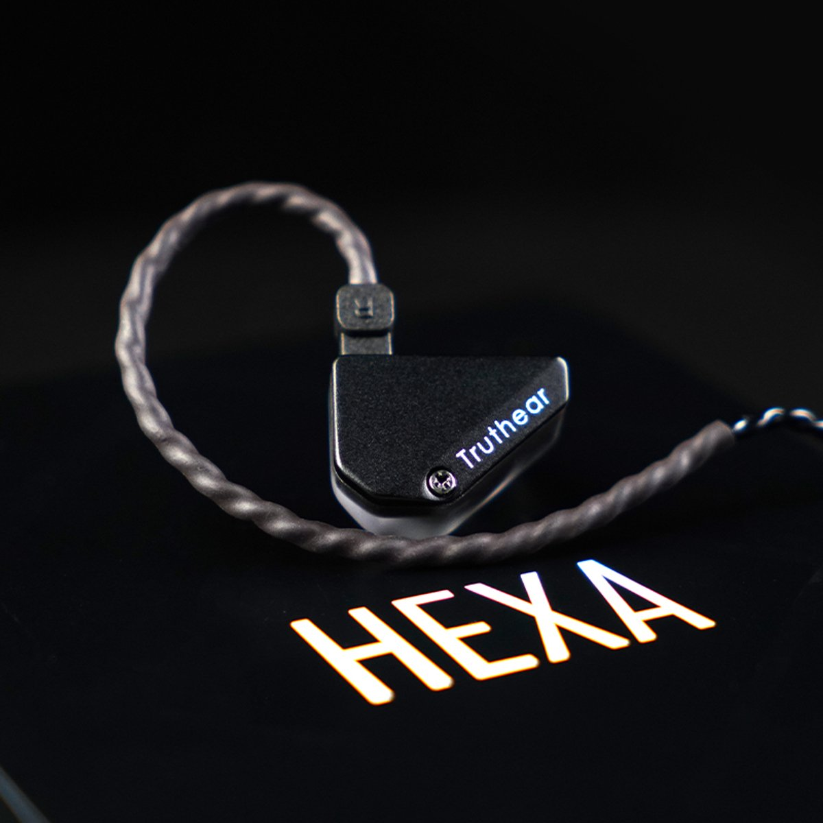 Schwarzer In-Ear-Monitor mit den Wörtern "Truthear" und "HEXA" auf einer schwarzen Oberfläche. Graues Kabel verbindet den Kopfhörer.
