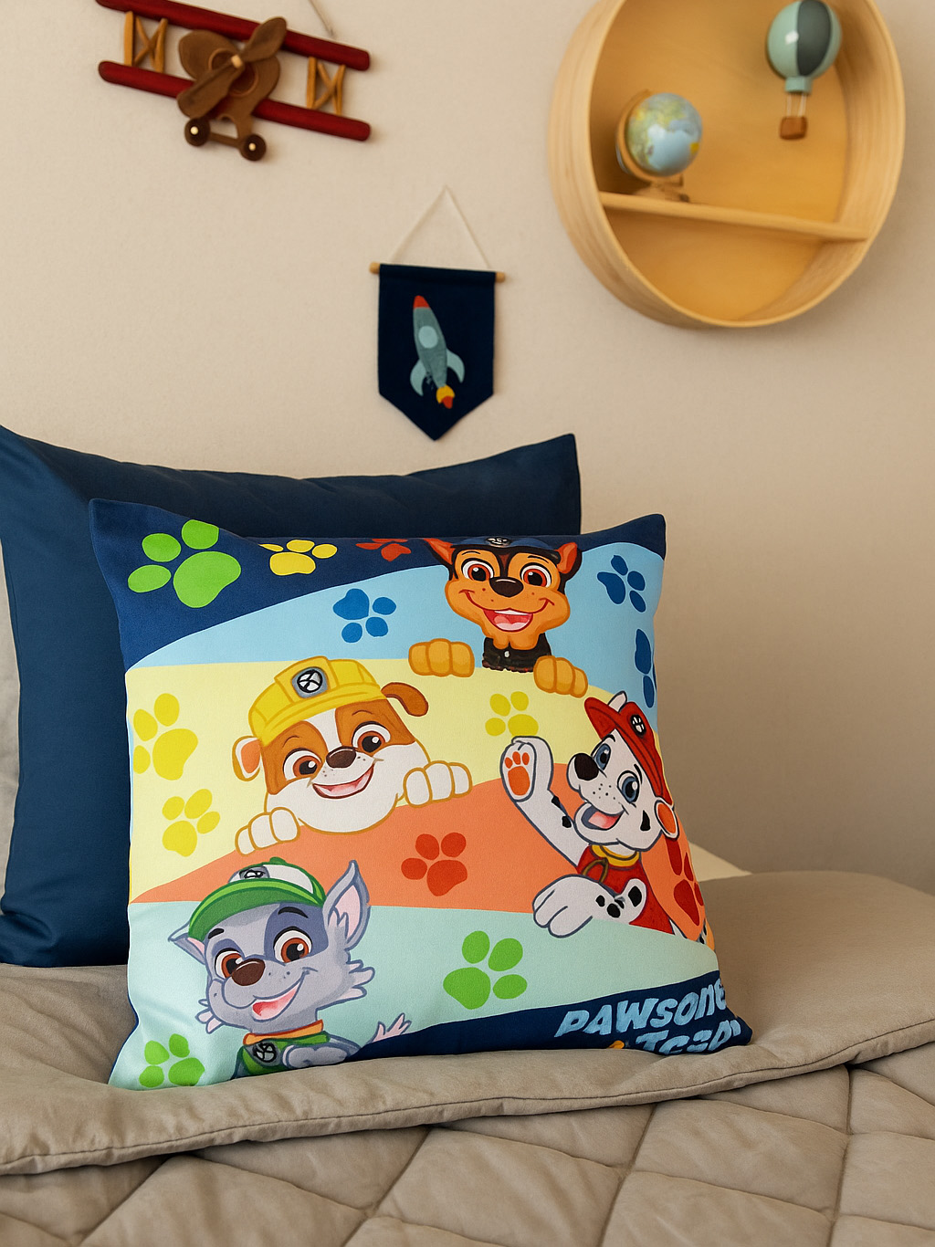 Ein Kissen mit Paw Patrol-Charakteren liegt auf einem Bett. Dekorationen sind ein Flugzeug und ein Regal mit einem Globus.
