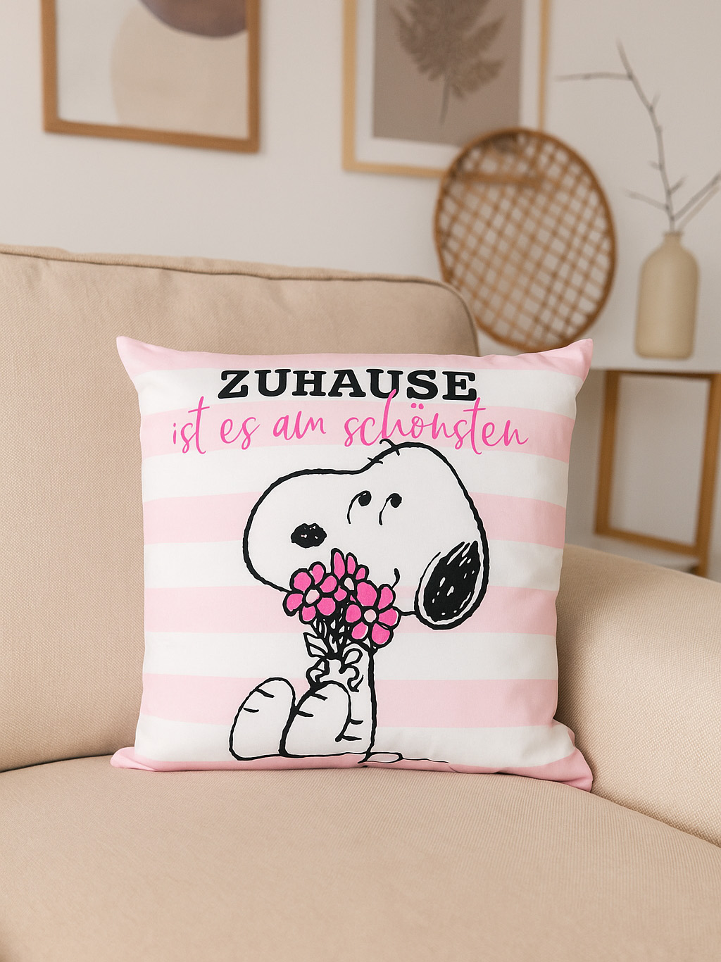Ein Snoopy-Kissen mit rosa Streifen und Blumen auf einem beigen Sofa. Auf dem Kissen steht 'Zuhause ist es am schönsten'.