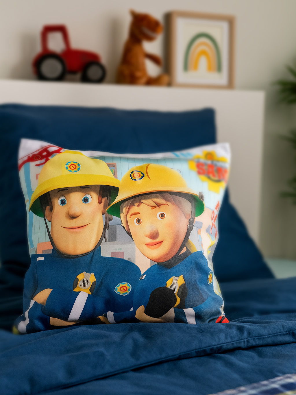 Kissen mit zwei Cartoon-Feuerwehrleuten in Uniform. Spielzeug und ein gerahmtes Regenbogenkunstwerk im verschwommenen Hintergrund.