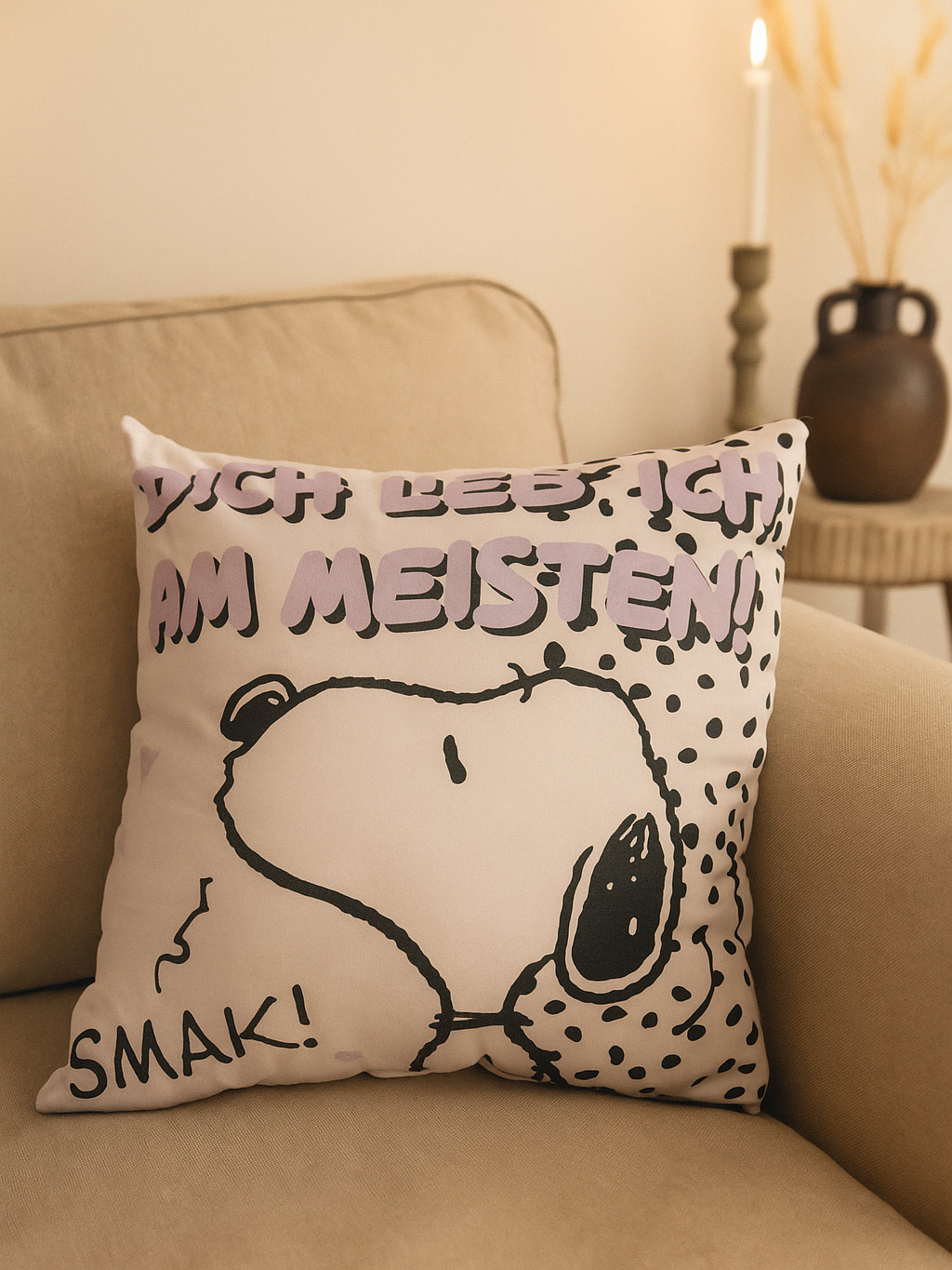 Ein Kissen mit Snoopy darauf, auf einem beigen Sofa platziert. Das Kissen hat lila Text, und im Hintergrund brennt eine Kerze.