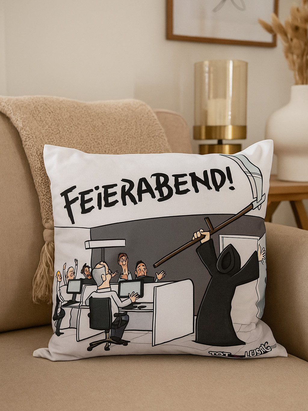 Kissen mit Cartoon von Büroangestellten und dem Sensenmann. Text lautet 'Feierabend!'. Kissen auf einem beigen Sofa, neutraler Hintergrund.
