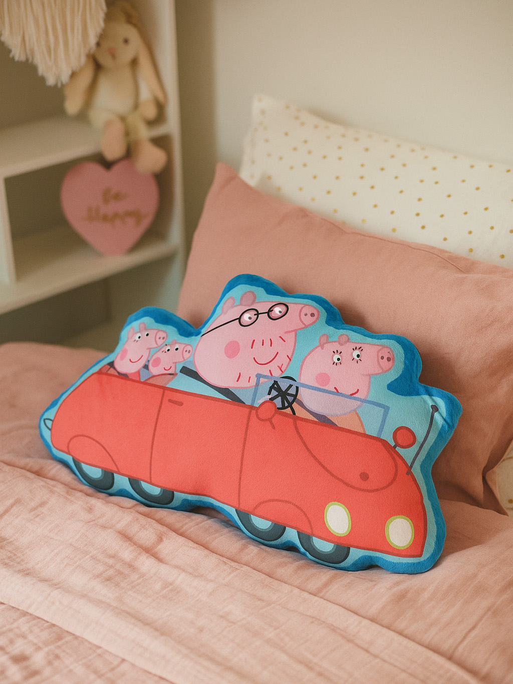 Ein Peppa Wutz-Autokissen auf einem rosa Bett. Im Hintergrund befinden sich weitere Kissen und ein Regal mit Dekoration.