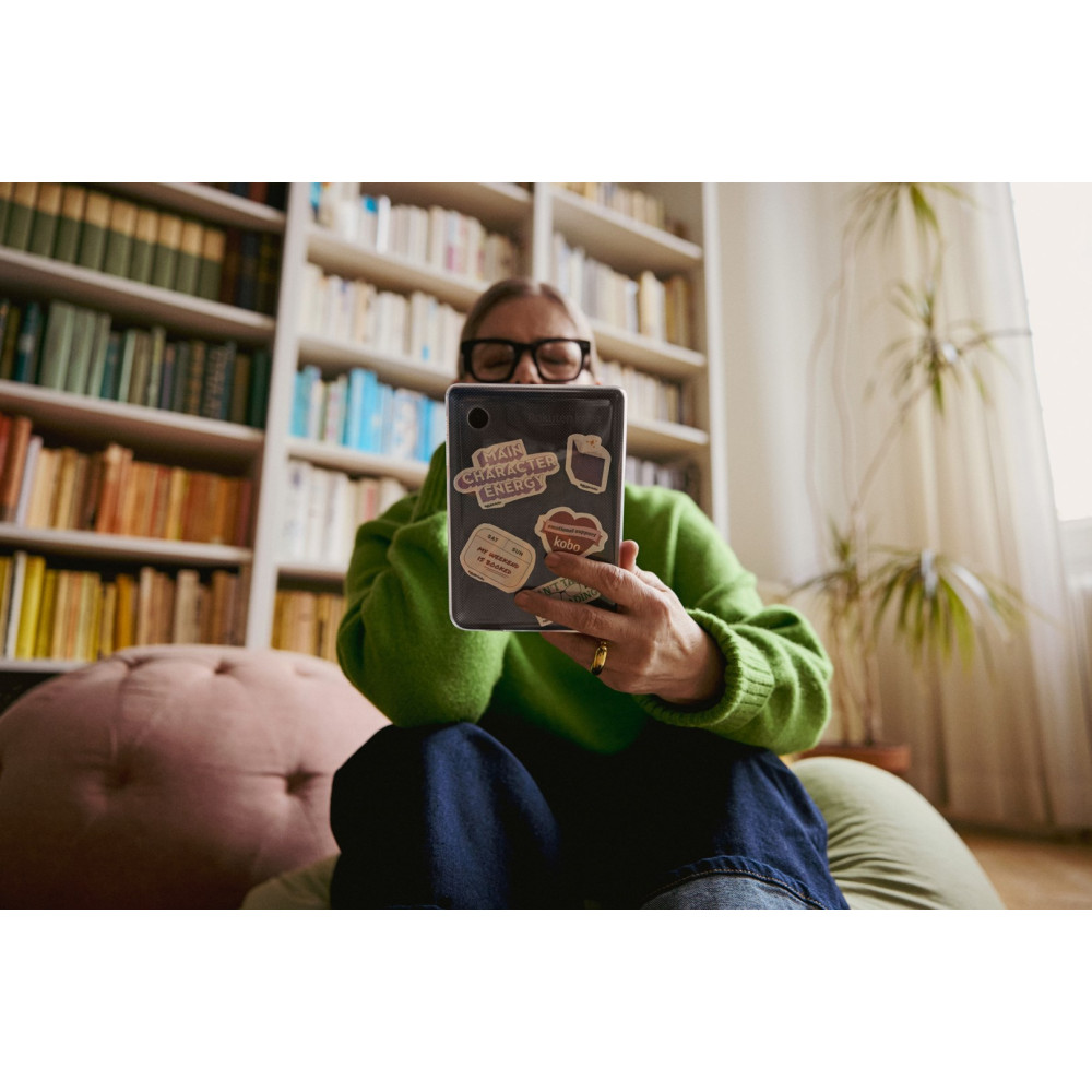 Osoba w okularach trzyma przed sobą tablet z naklejkami. W tle regały z książkami. Ma na sobie zielony sweter.