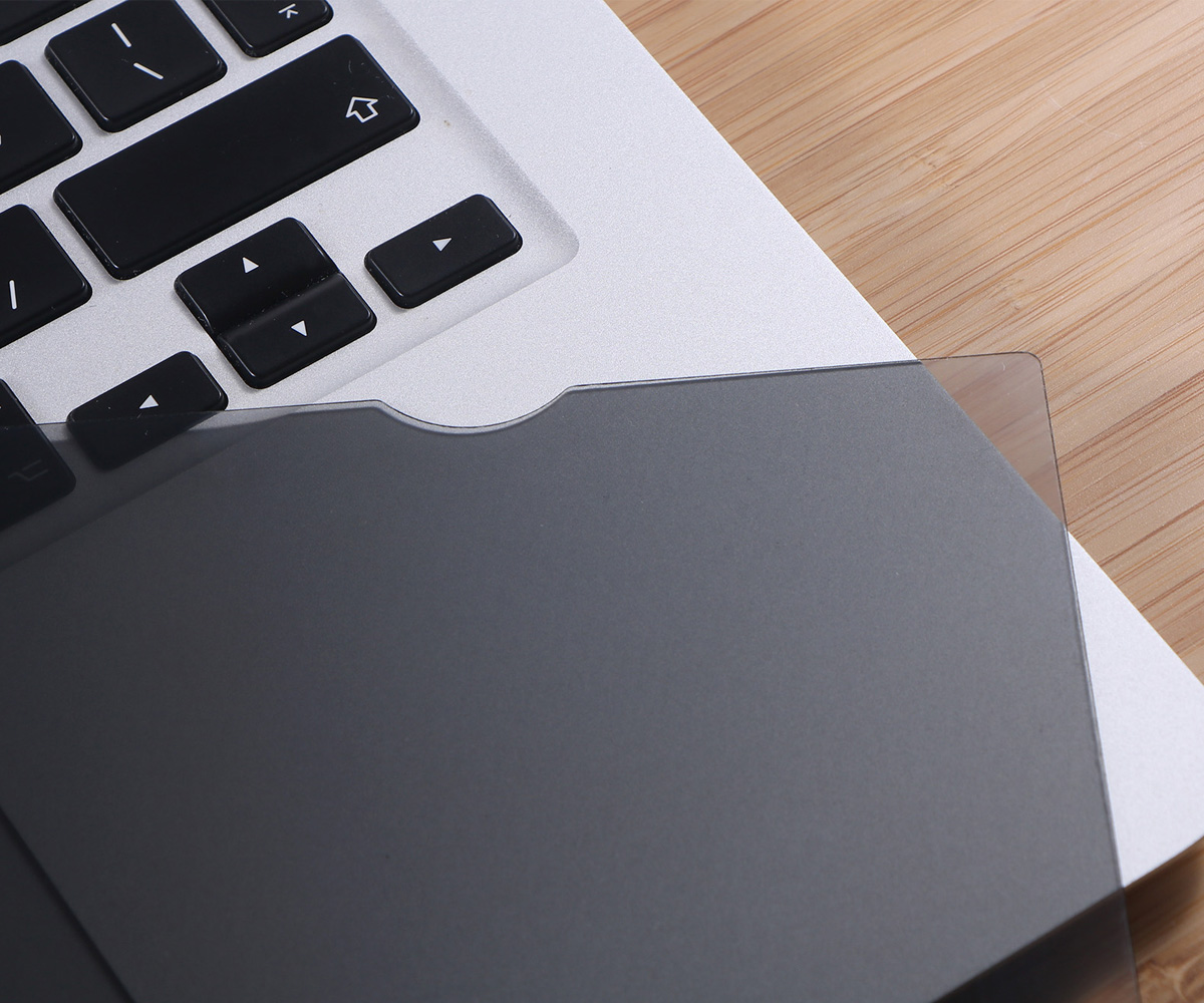 Klawiatura laptopa z nakładką na ekran chroniącą prywatność. Klawiatura jest srebrna z czarnymi klawiszami. W tle drewniany stół.