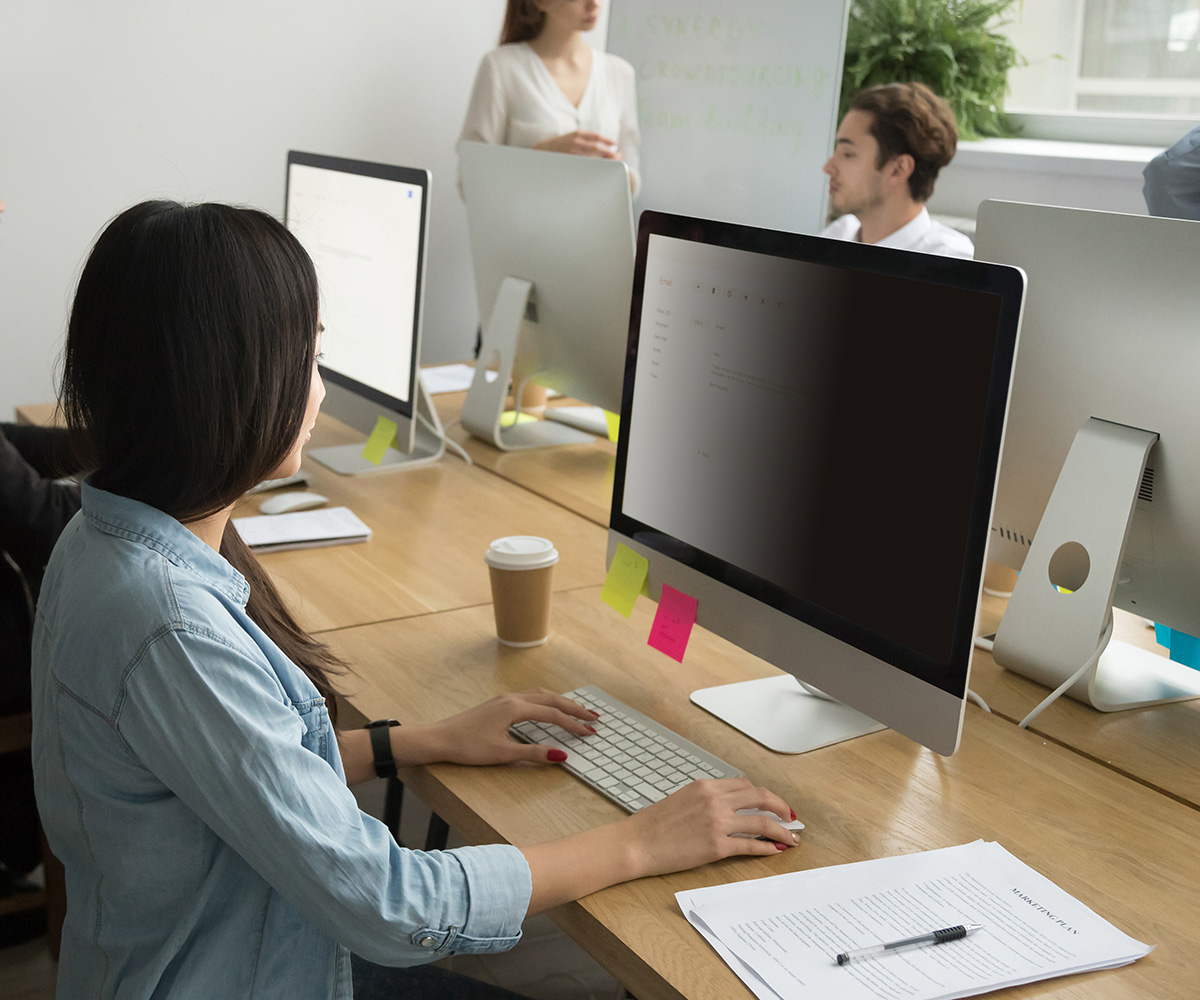 Kobieta w niebieskiej koszuli pracuje na komputerze. Otoczenie biurowe z innymi ludźmi, monitorami, papierami i filiżanką kawy.