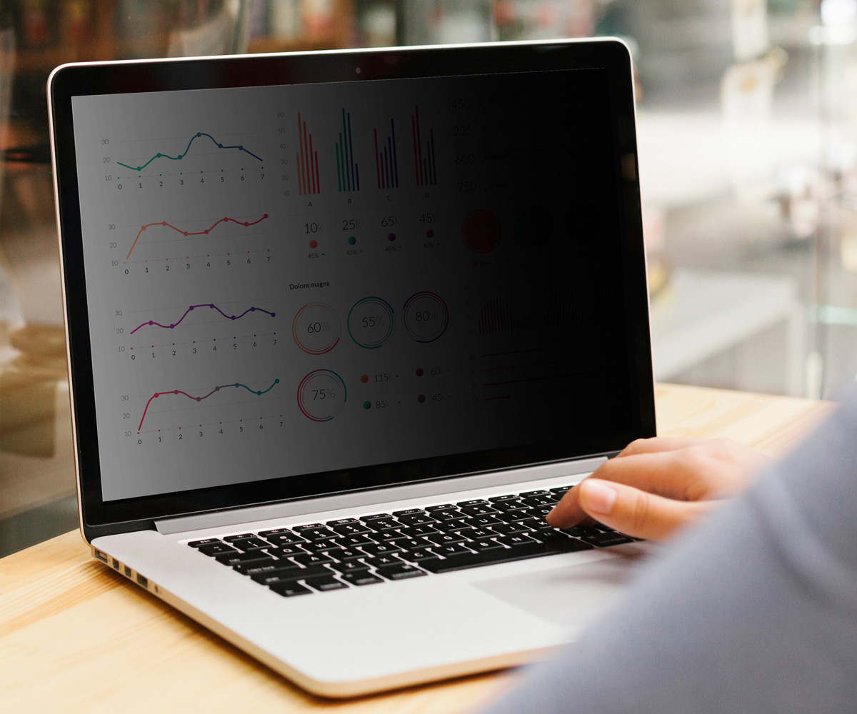 Laptop wyświetla wykresy i dane. Ręka osoby pisze na klawiaturze. Tło jest rozmyte, pokazując stół i okno.