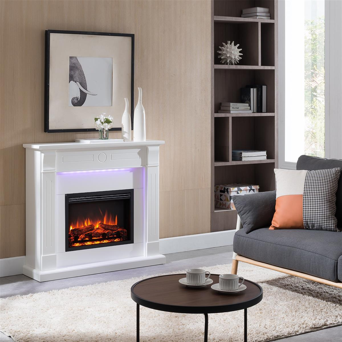 Wohnzimmer mit weißem Kamin, Elefantenkunst, grauem Sofa, rundem Tisch mit Tassen und einem Bücherregal. Der Kamin hat einen Feuereffekt.