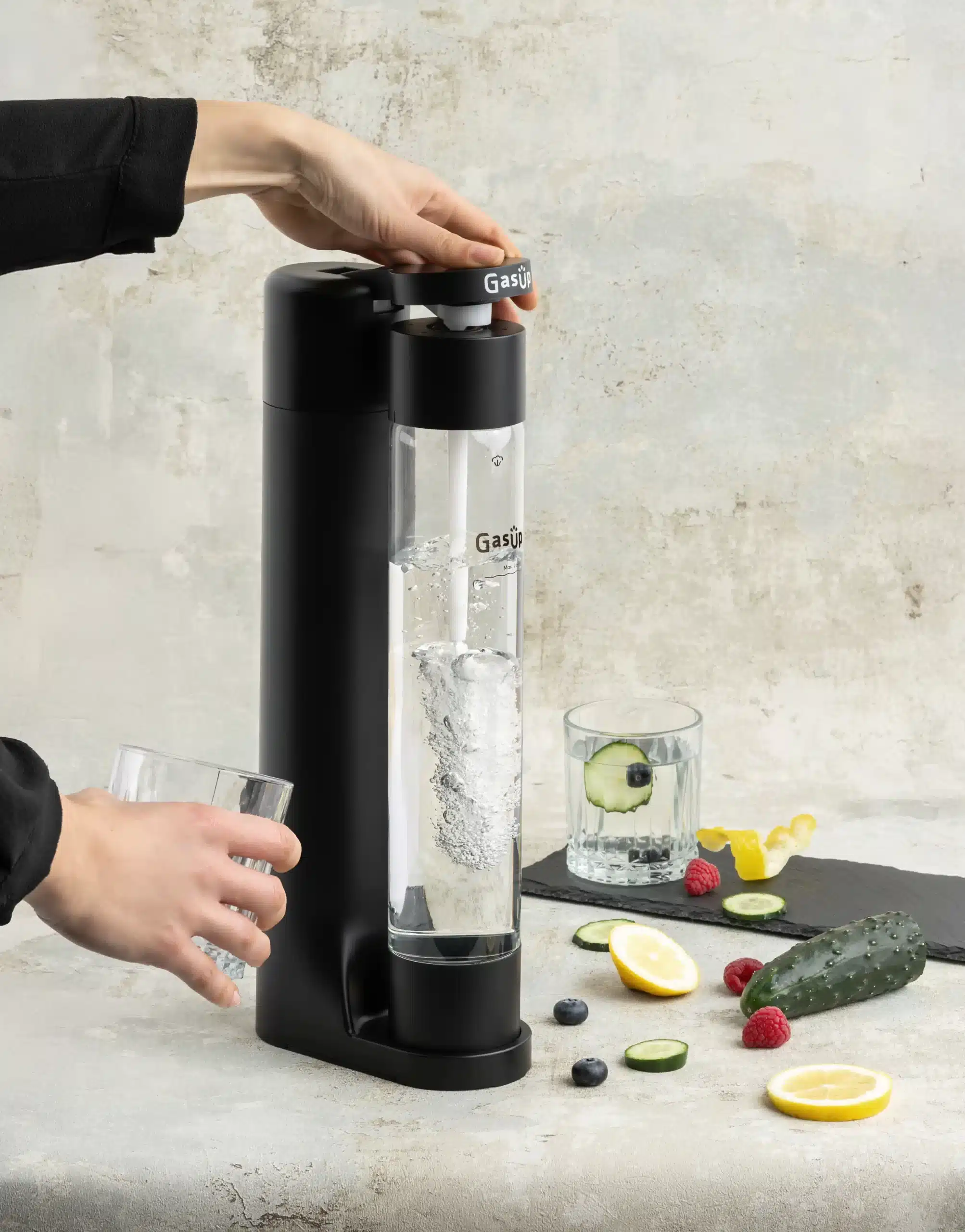 Una persona usa un produttore di soda. Un bicchiere d'acqua, cetriolo e limone sono su una superficie.