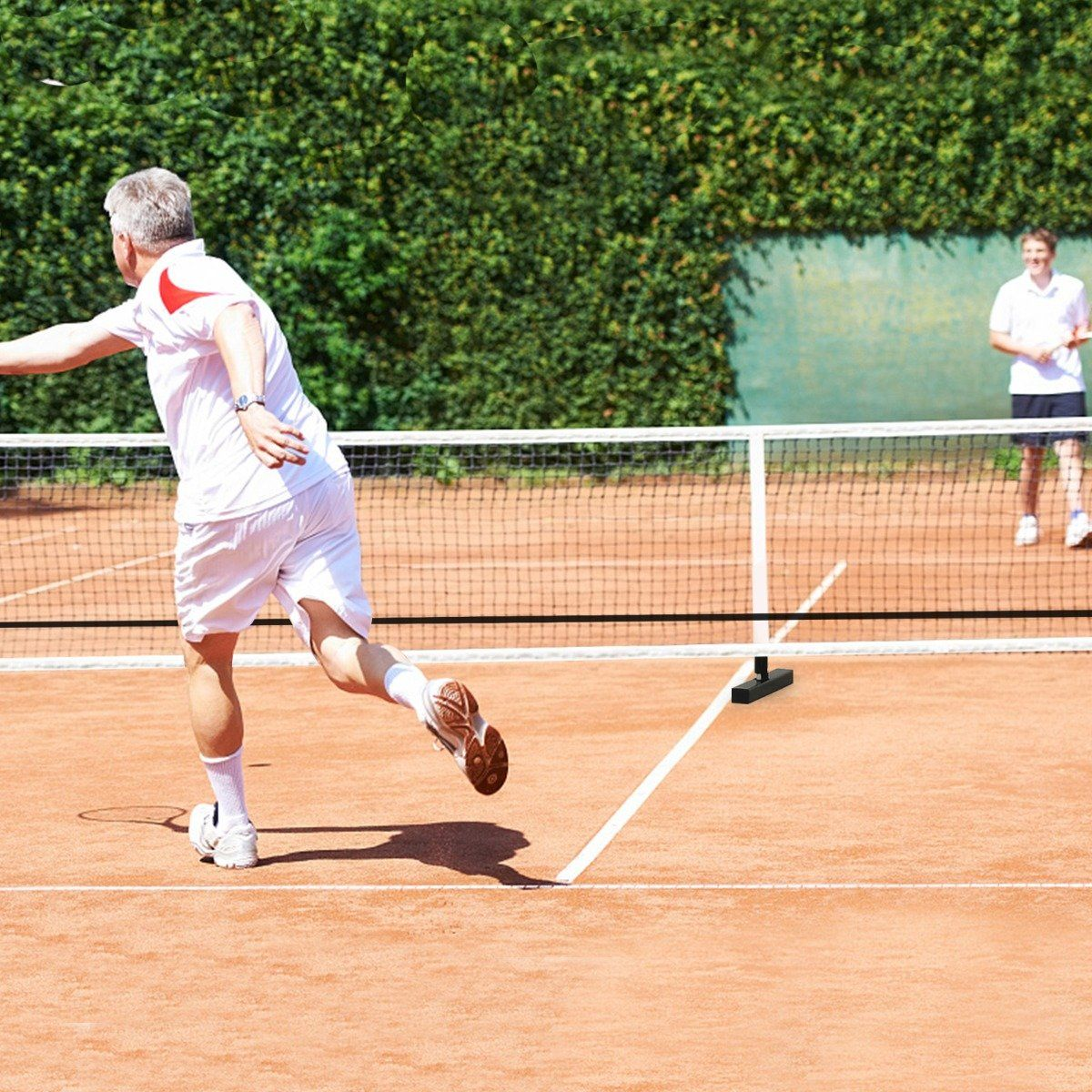 Un hombre de blanco juega al tenis, golpeando una pelota. Otro hombre está en la red. Fondo de seto verde. Pista naranja.