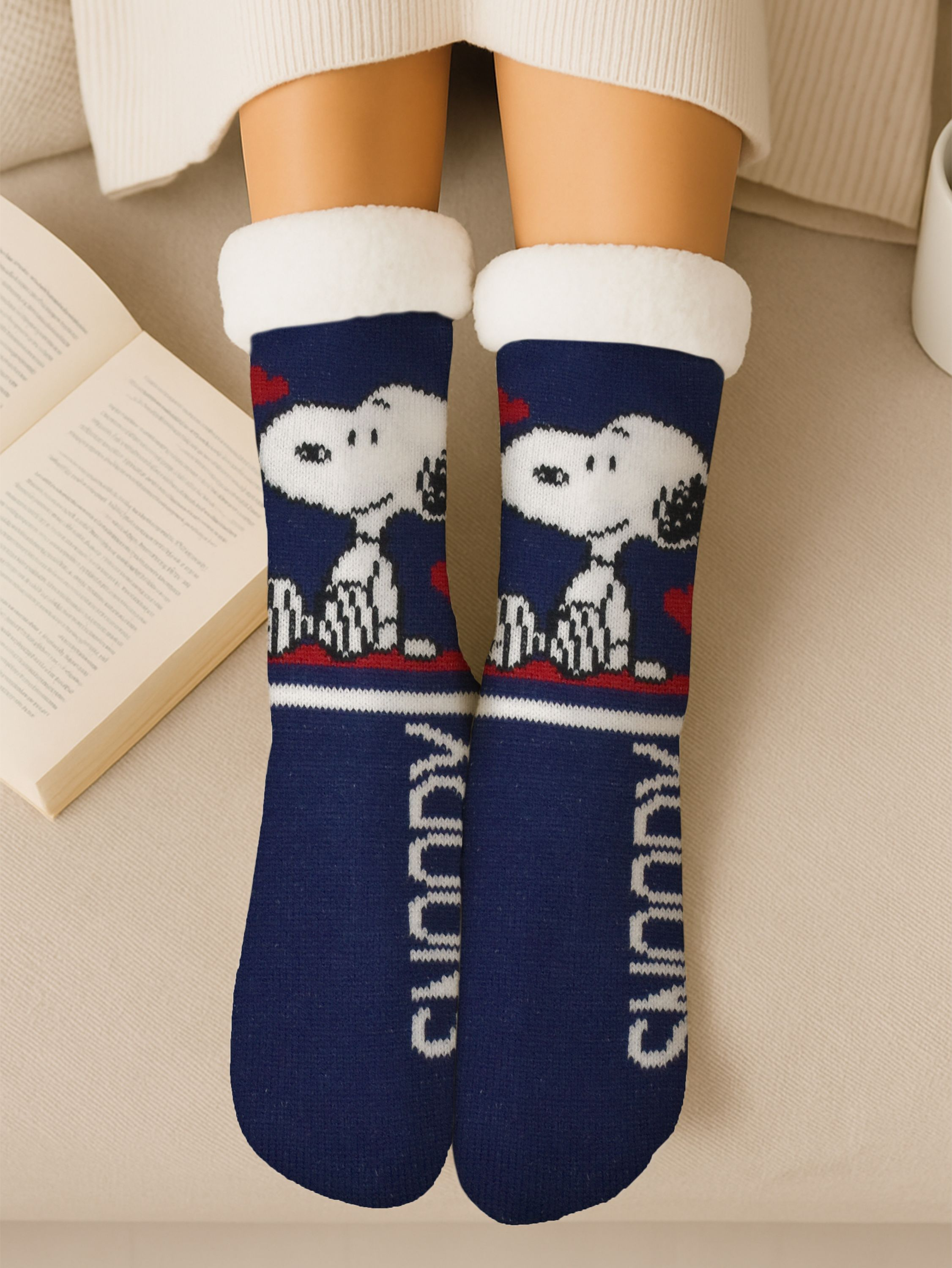 Beine einer Person mit marineblauen Snoopy-Socken mit weißer Bordüre und Sherpa-Futter. Offenes Buch links, beiger Hintergrund.