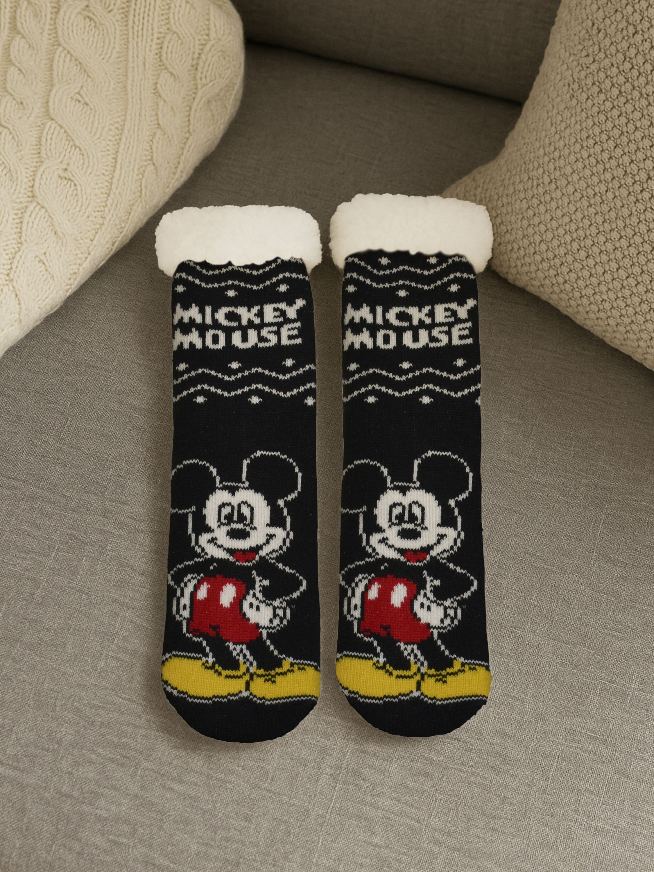 Zwei schwarze Socken mit Mickey Mouse-Design. Weiße Manschetten, rote Shorts und gelbe Schuhe. Beige Kissen im Hintergrund.