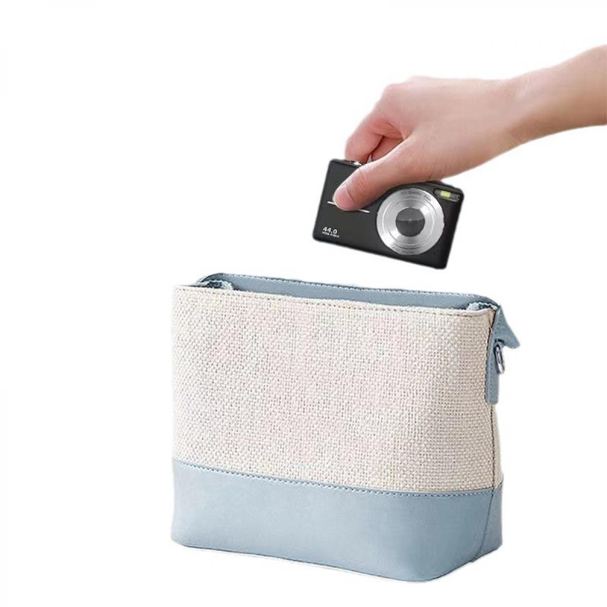 Hand legt eine schwarze Digitalkamera in eine weiß-hellblaue Tasche. Die Kamera hat ein silbernes Objektiv.