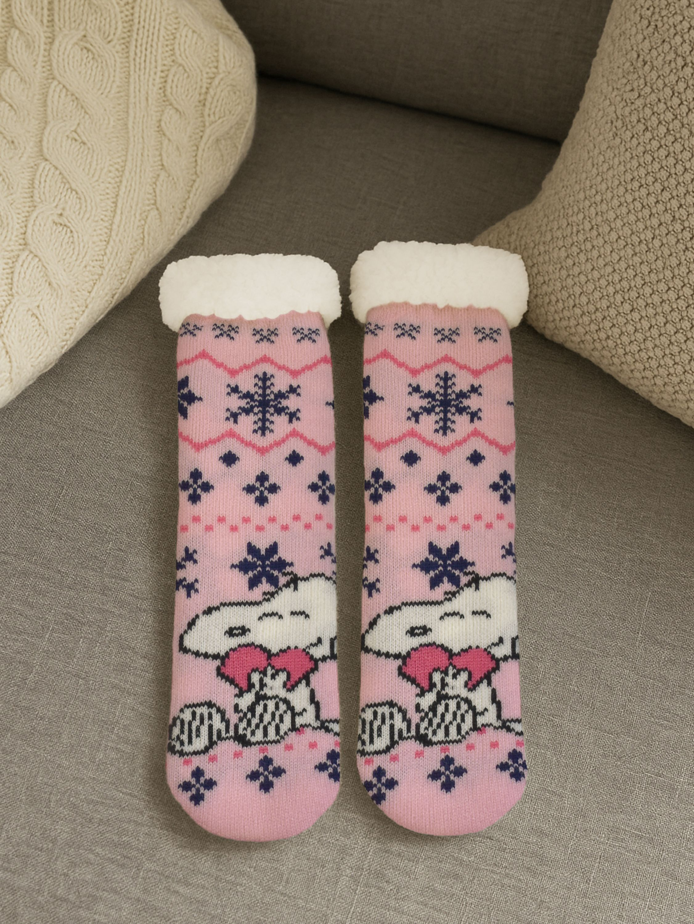 Zwei rosa Socken mit Snoopy-Design und Schneeflocken. Sie haben weiße, flauschige Oberteile, auf einem grauen Sofa mit Kissen.