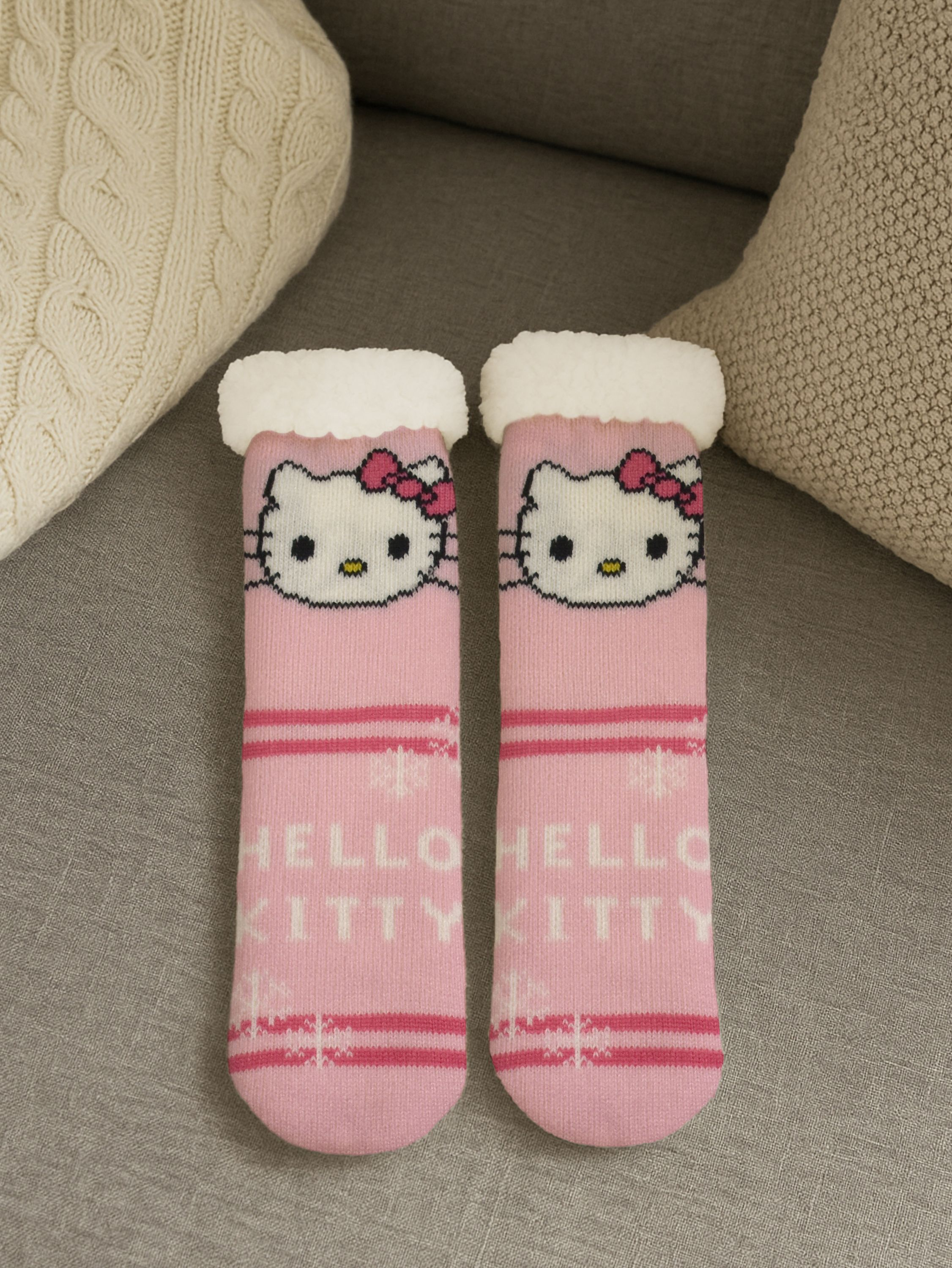 Paar rosa Hello Kitty Socken mit weißem Besatz auf einem grauen Sofa mit Kissen.