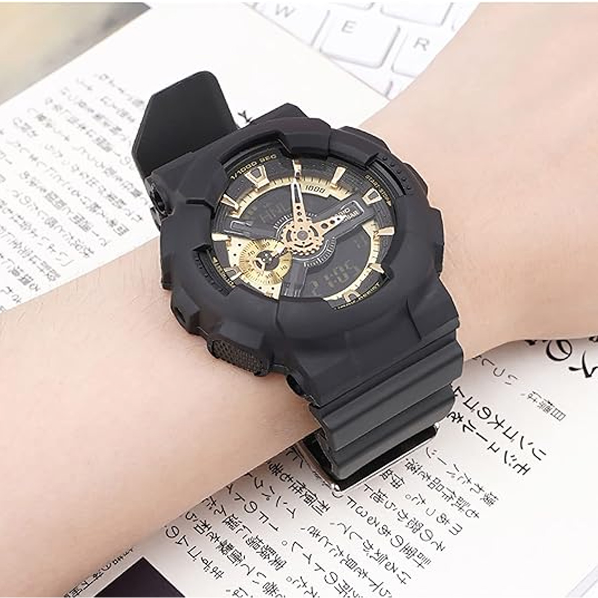 Eine schwarze Armbanduhr mit goldenem Zifferblatt liegt auf einem Handgelenk, über einem Buch mit japanischem Text und einer Tastatur im Hintergrund.