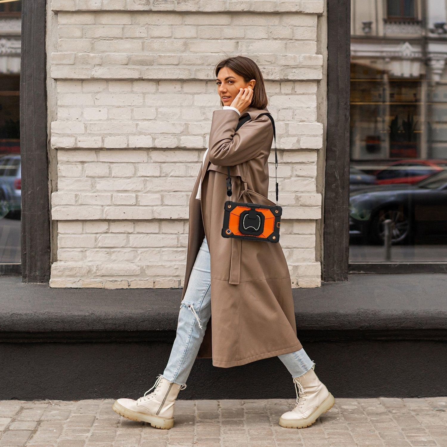 Eine Frau in einem langen Mantel und Jeans geht auf einer Straße. Sie trägt eine Tasche mit orangefarbener Hülle. Weiße Stiefel.