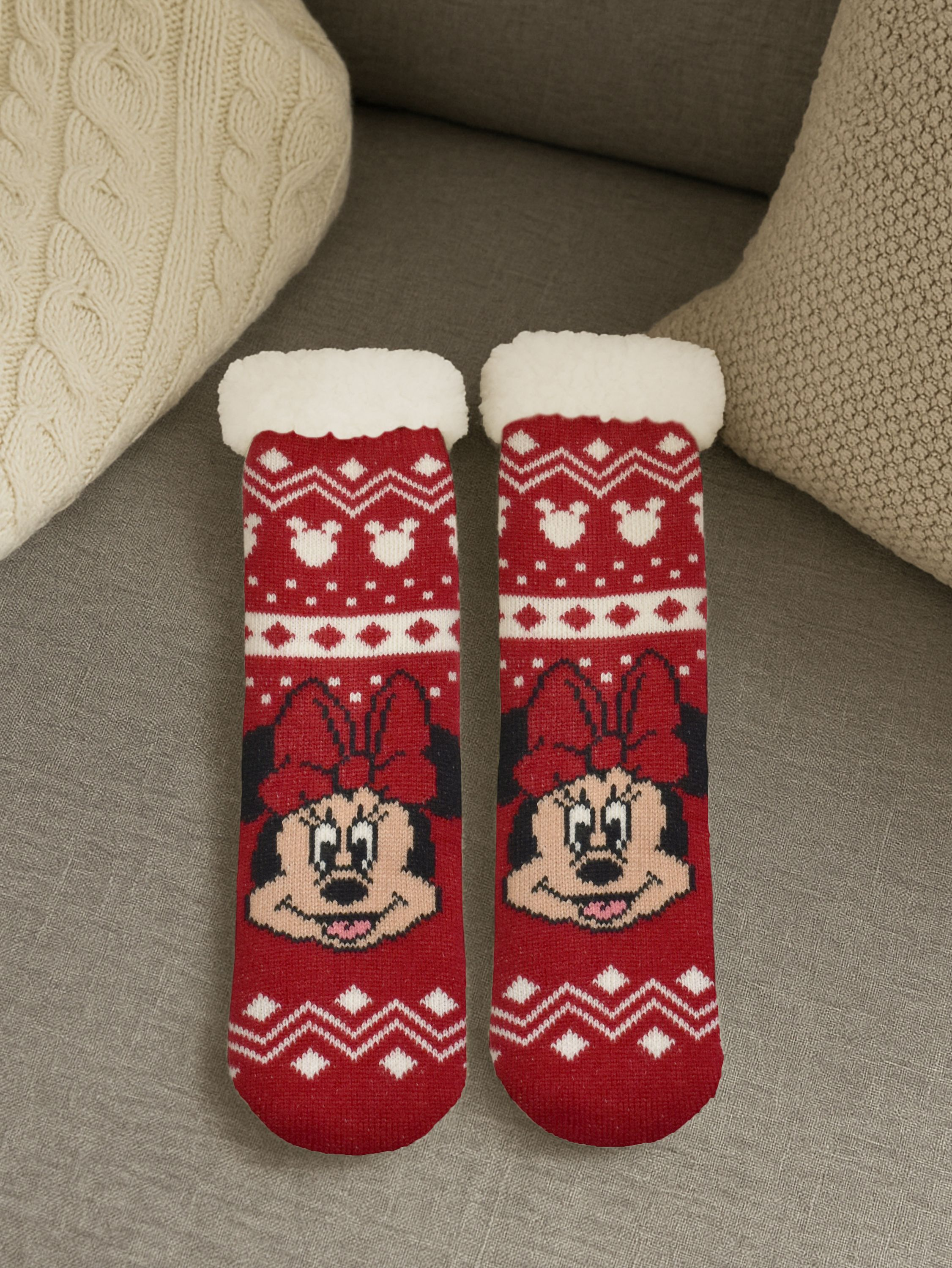 Zwei rote Socken mit Minnie-Maus-Gesichtern, weißem Besatz und einem dekorativen Muster auf einer grauen Couch.