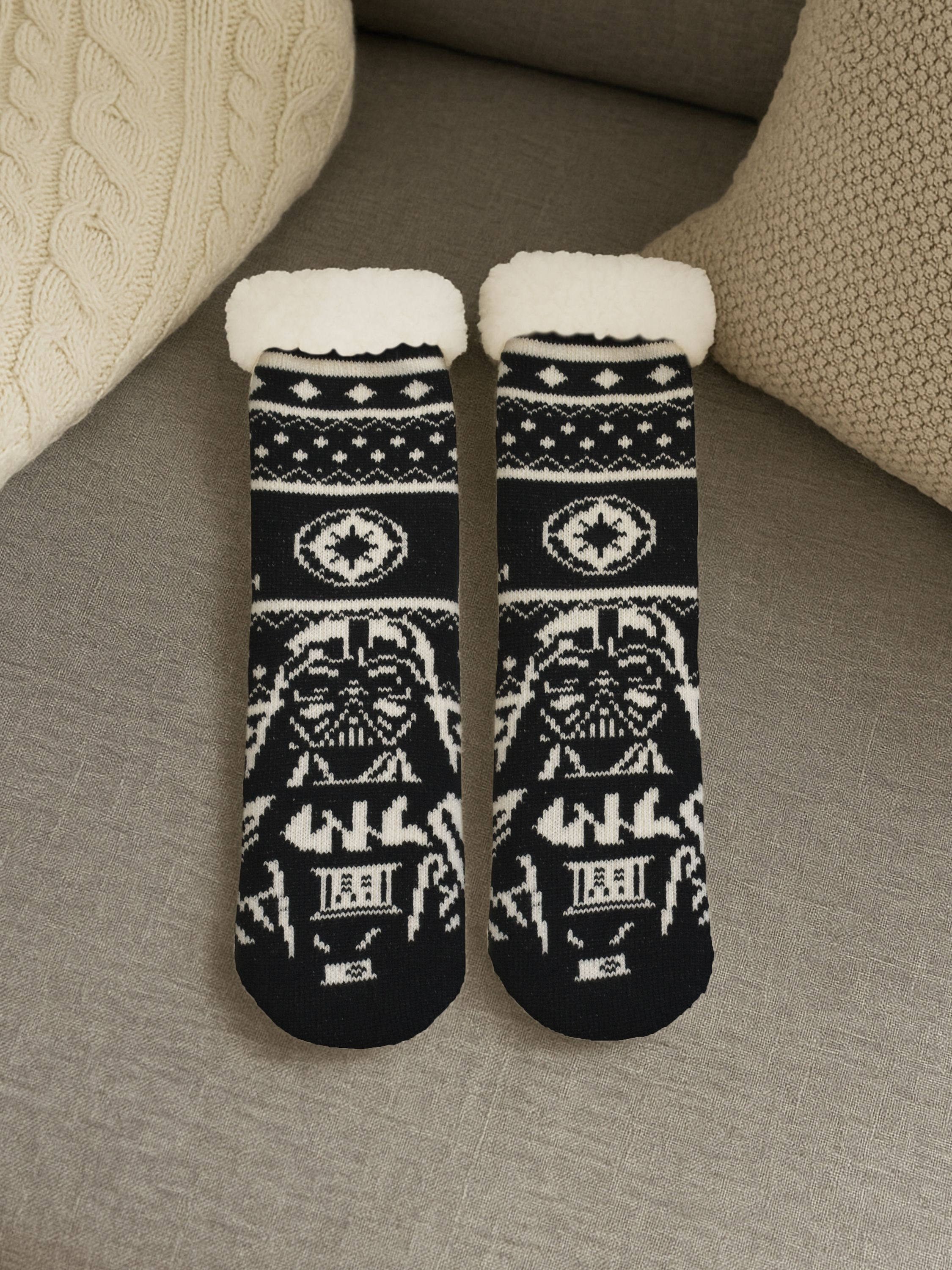 Paar schwarze Socken mit weißem Darth Vader-Design, flauschige weiße Oberteile, auf grauer Oberfläche mit Kissen.